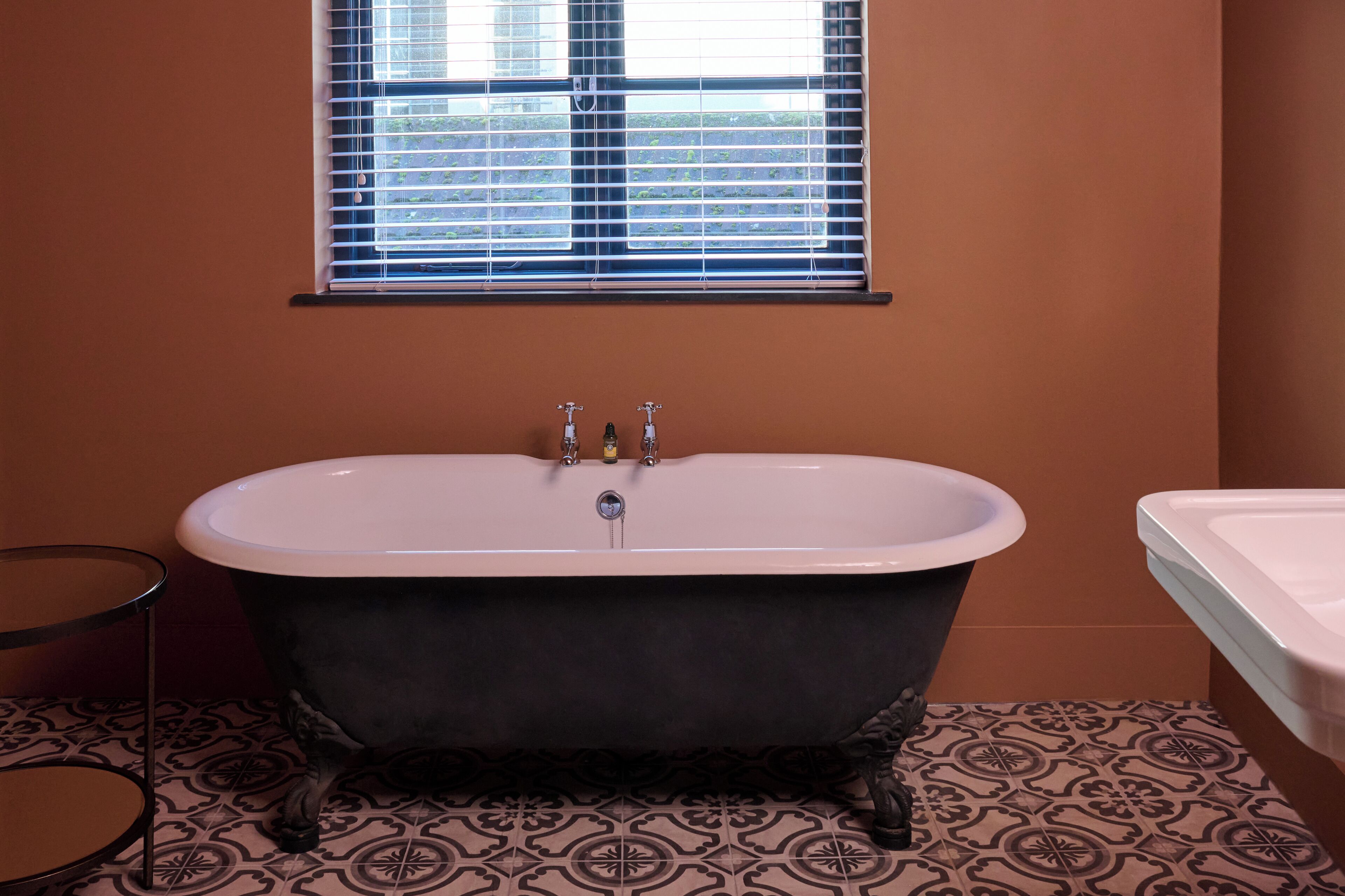 A bathroom with a freestanding bathtub, a window with blinds, and patterned floor tiles.