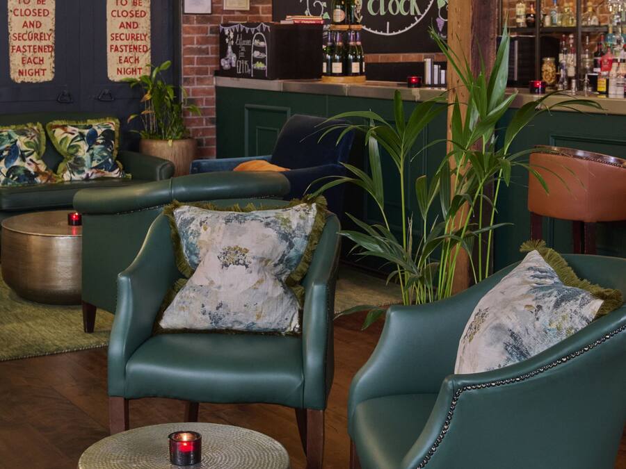 En hyggelig bar med grønne læderstole, et rundt bord med et stearinlys og en murstensvæg med et ur og barskabe.