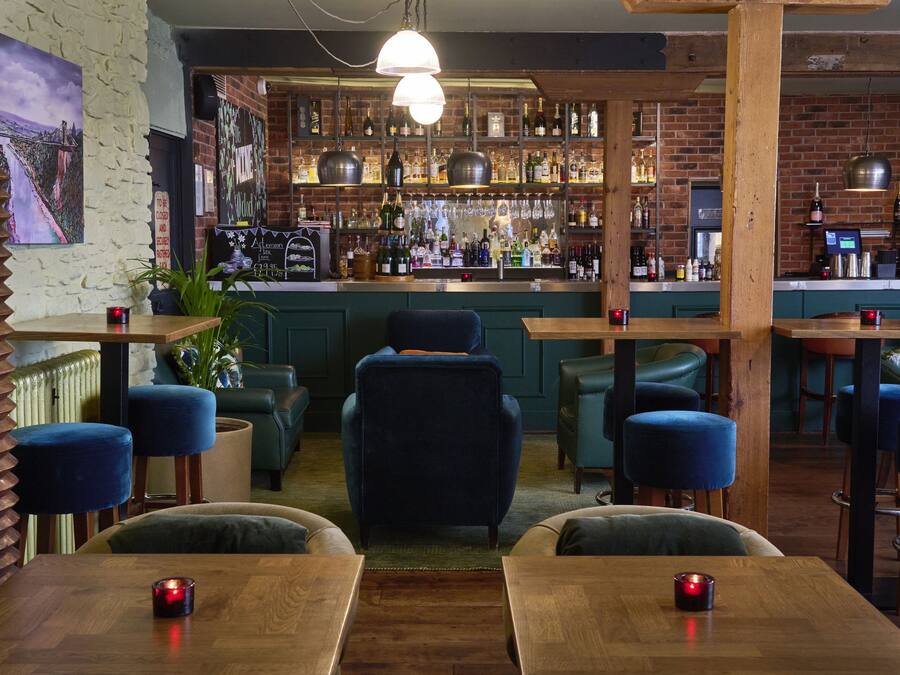 A bar area with a brick wall, shelves of bottles, and a counter. There are blue chairs and tables with candles.