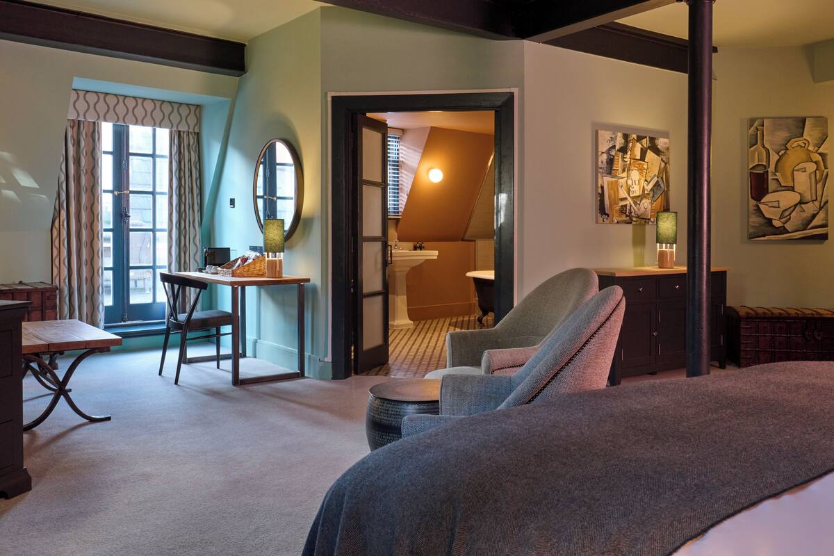 Ein Hotelzimmer mit Bett, Schreibtisch, Stuhl und einem kleinen Tisch. Es gibt ein Fenster mit Vorhängen, ein Bild an der Wand und eine Tür zu einem weiteren Raum.