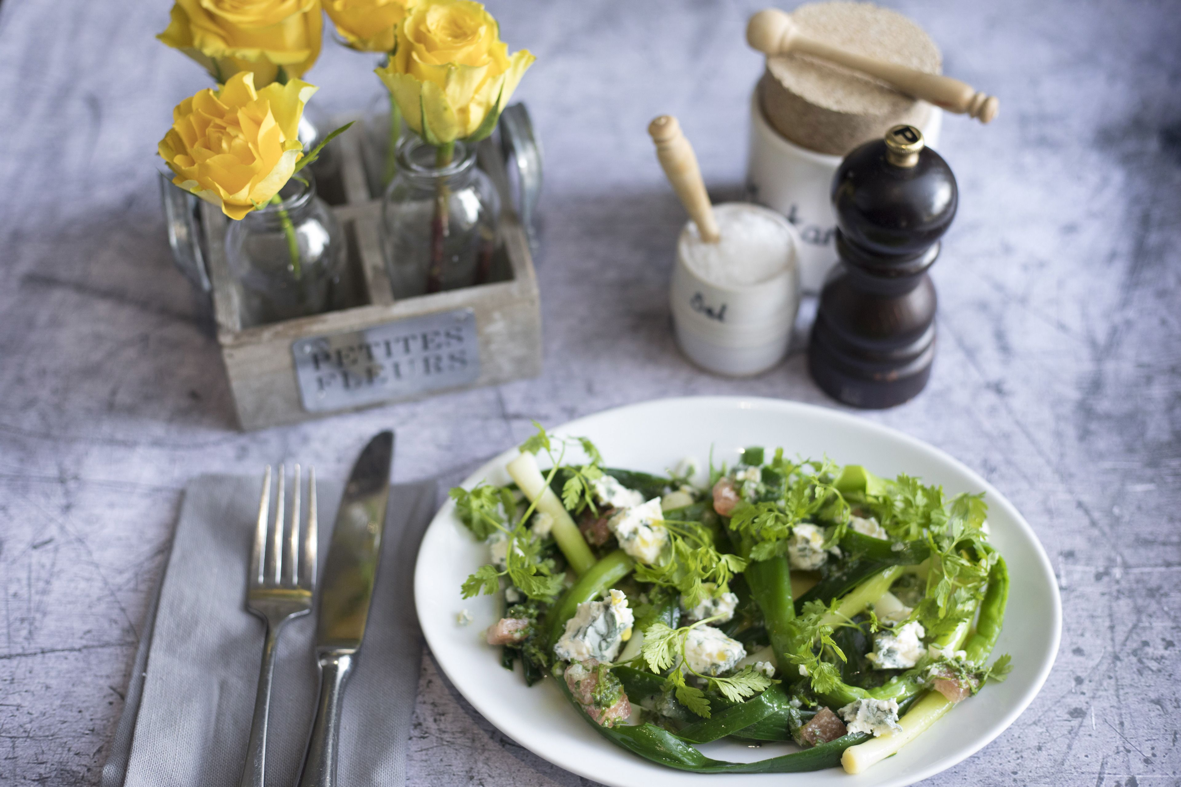 Una ensalada con vegetales verdes y queso feta, un cuchillo y un tenedor sobre una servilleta, y un jarrón con flores amarillas al fondo.