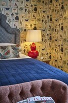 A bedroom with a blue quilted bedspread, a red lamp, and a wall decorated with botanical prints.