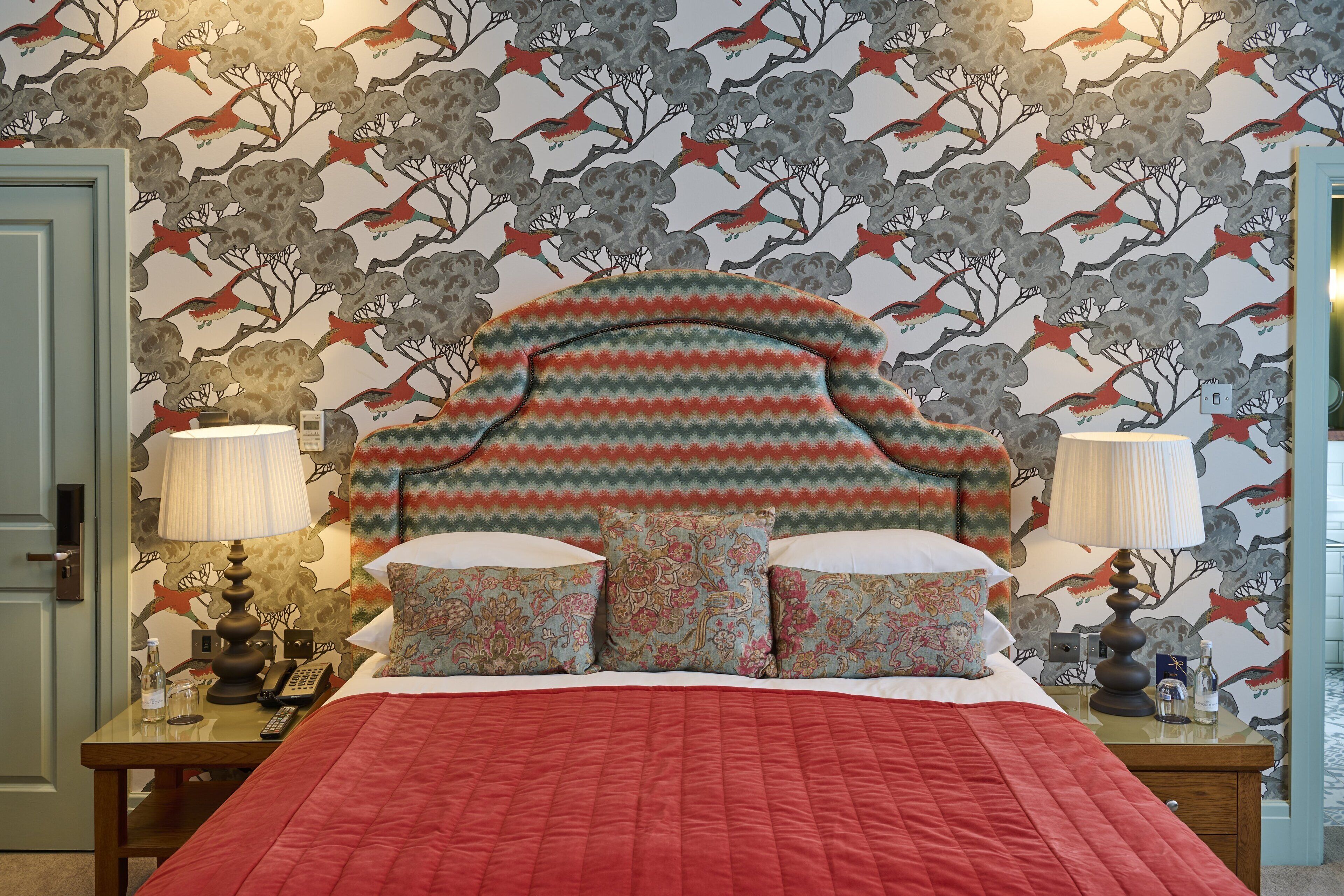 A hotel room with a patterned wallpaper, a red quilted bedspread, and floral-patterned pillows.