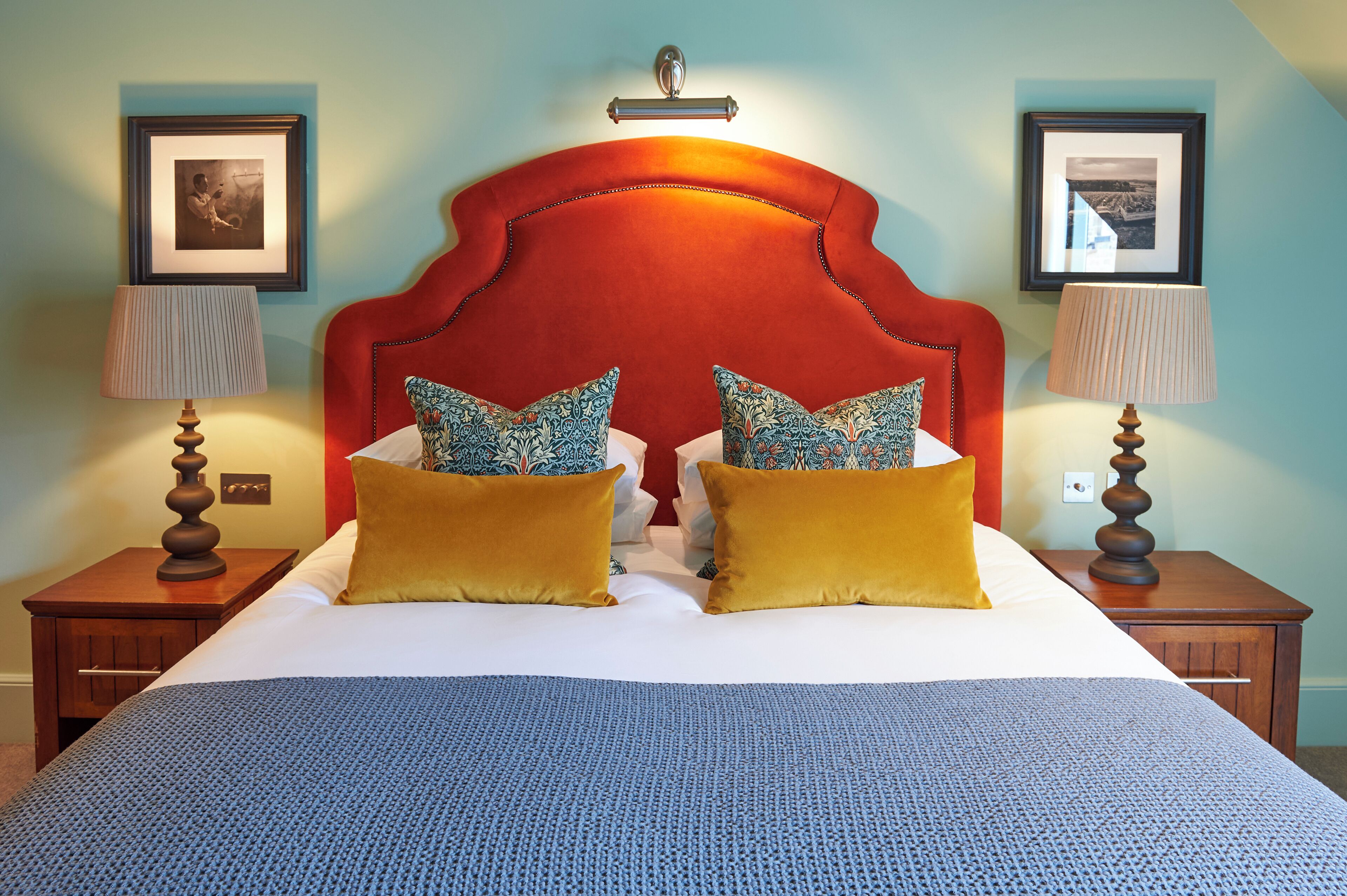 Un lit bien fait, avec une tête de lit rouge, deux coussins à motifs et une couverture bleue. Deux tables de chevet sont placées de chaque côté du lit, et des lampes ainsi que des tableaux encadrés ornent les murs.