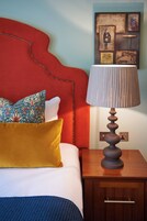 A neatly made bed with a red headboard, a mustard-colored pillow, and a blue patterned pillow. A wooden nightstand with a lamp is beside the bed.