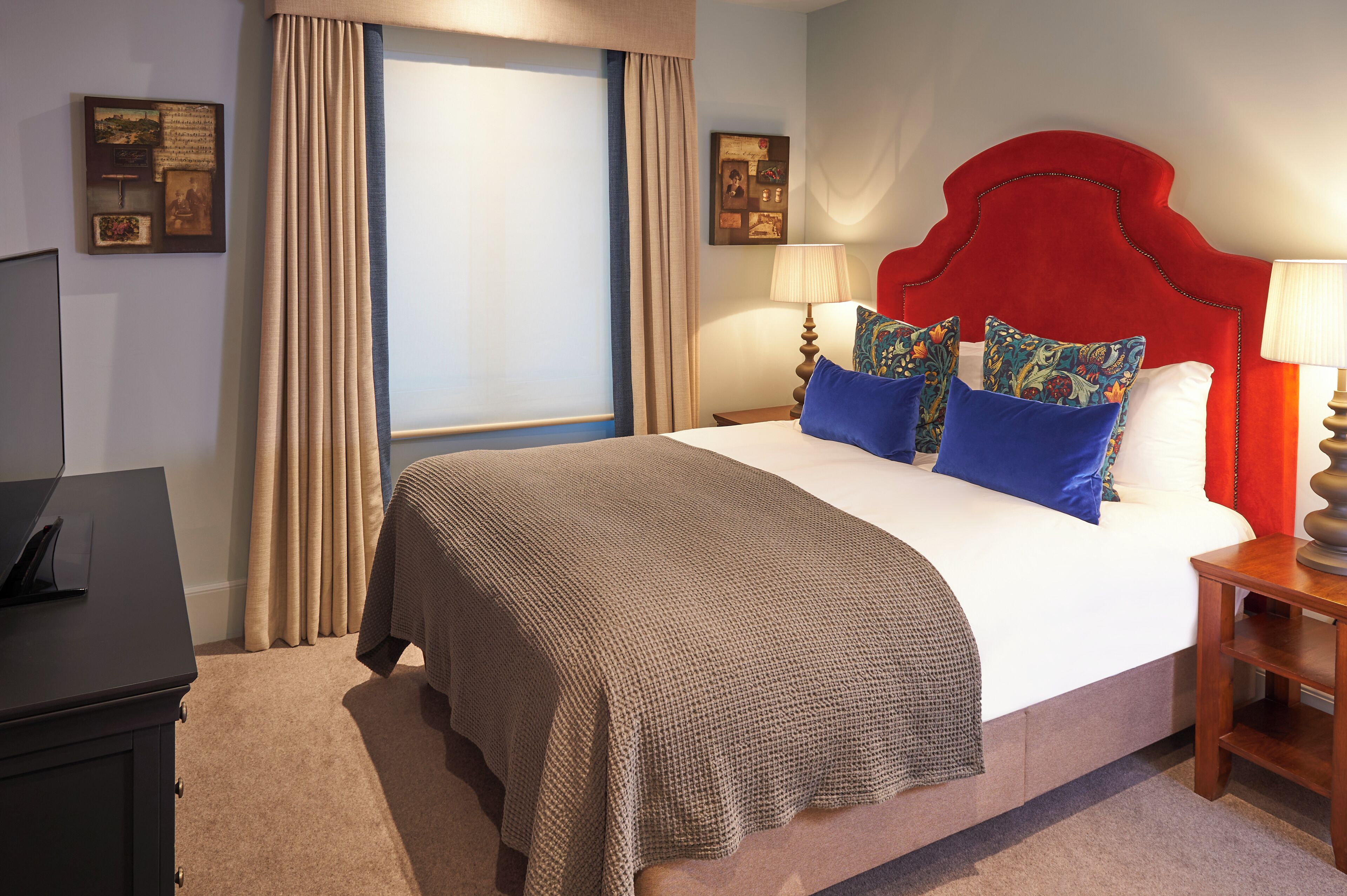 A neatly made bed with a red headboard, a bedside table with a lamp, and a window with curtains.