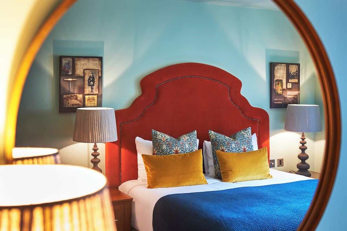 A hotel room with a red headboard, two lamps, a bed with a blue blanket, and two yellow pillows.
