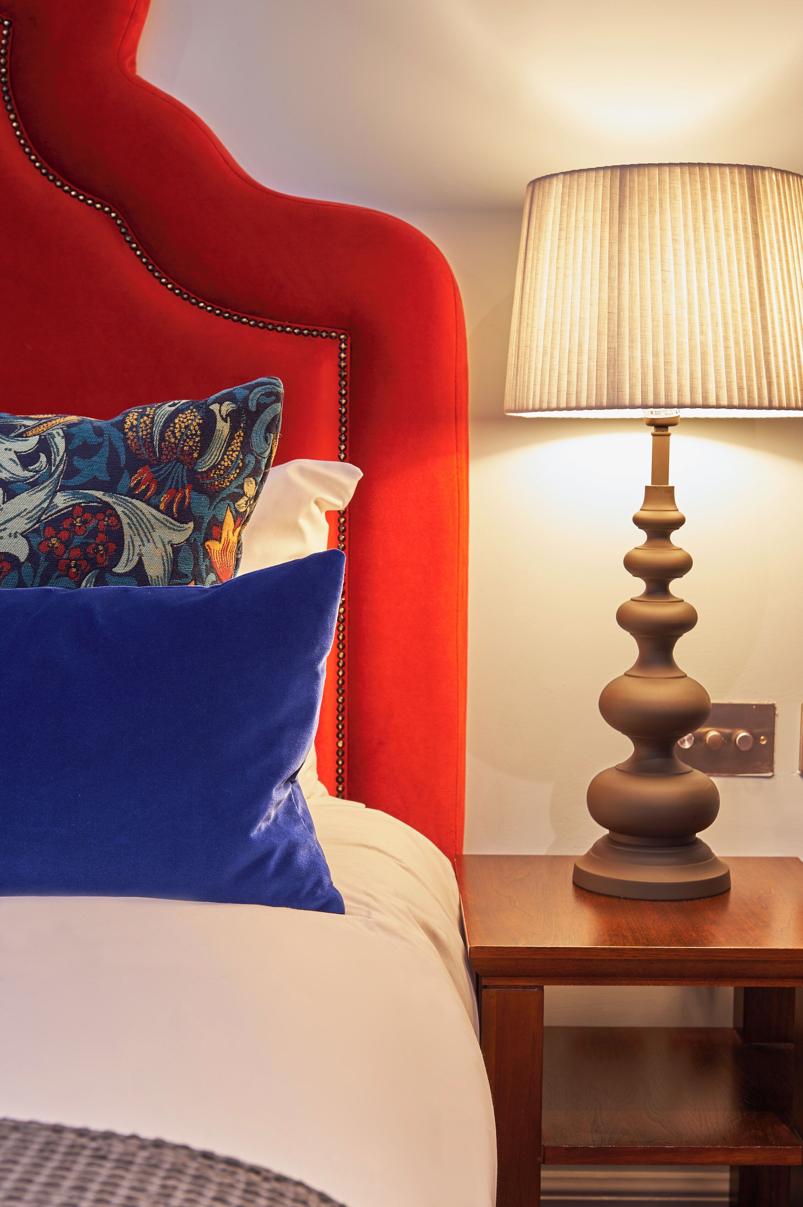 Een bed met een rode hoofdbord, blauw-witte kussens, en een houten nachtkastje met een lamp.