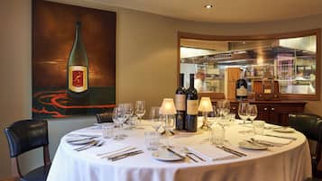 A dining table set with wine glasses, bottles, and cutlery, with a painting of a wine bottle on the wall.