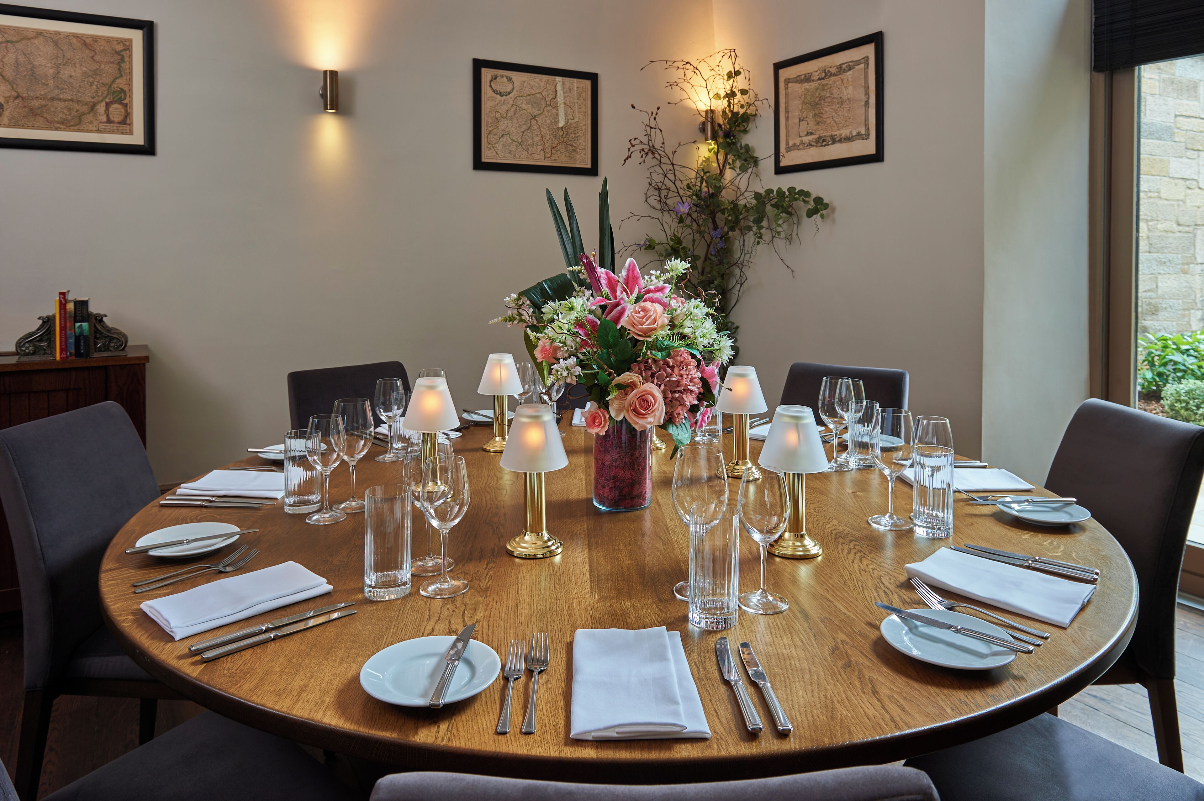 A round dining table set for six with white napkins, silverware, and clear glassware. A vase with pink flowers is centered on the table.