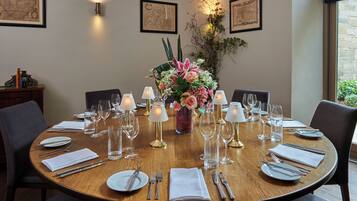 A round dining table set for six with white napkins, silverware, and clear glassware. A vase with pink flowers is centered on the table.