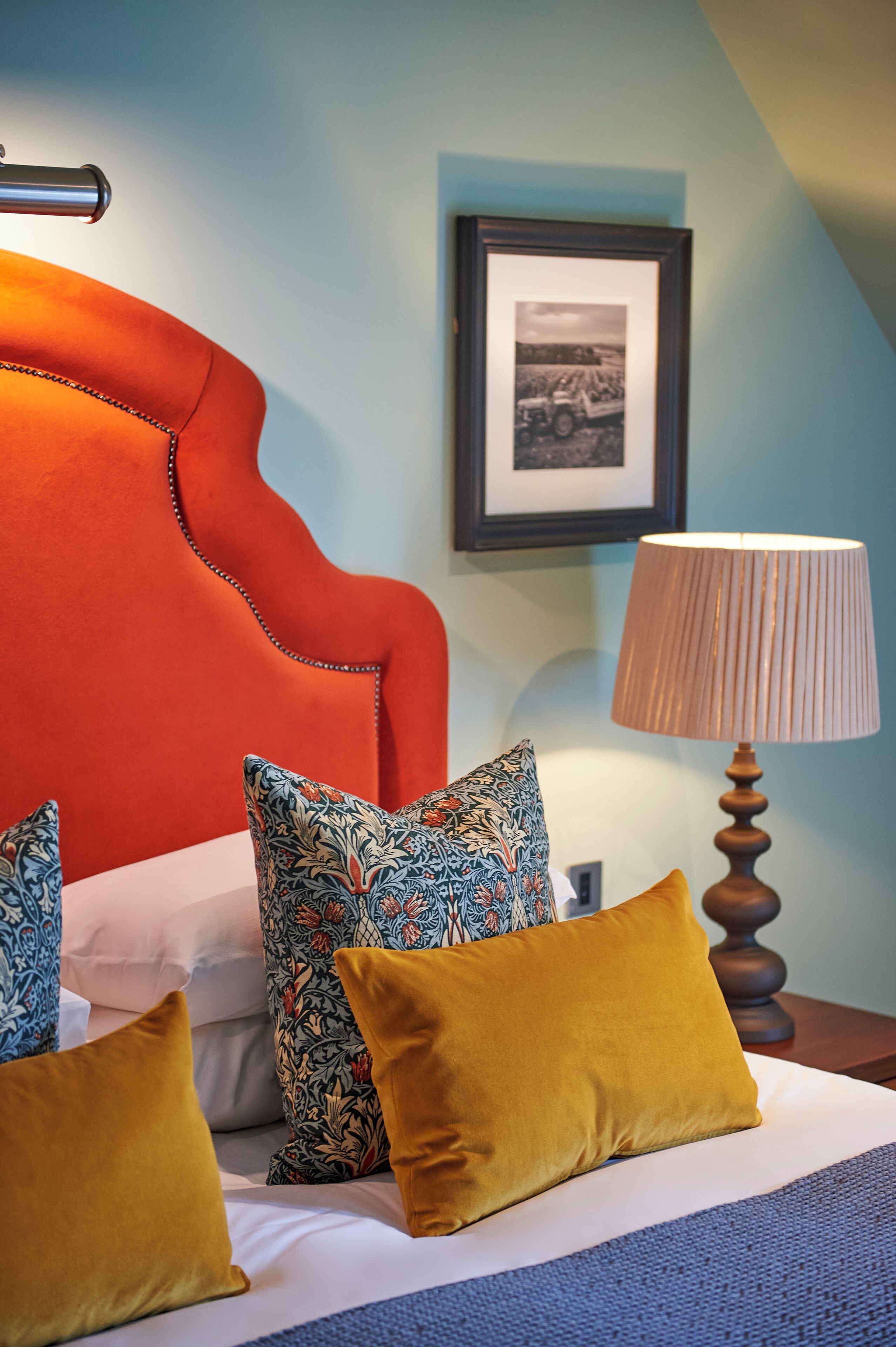A hotel room with a large orange headboard, patterned pillows, a wooden bedside table, and a framed picture on the wall.