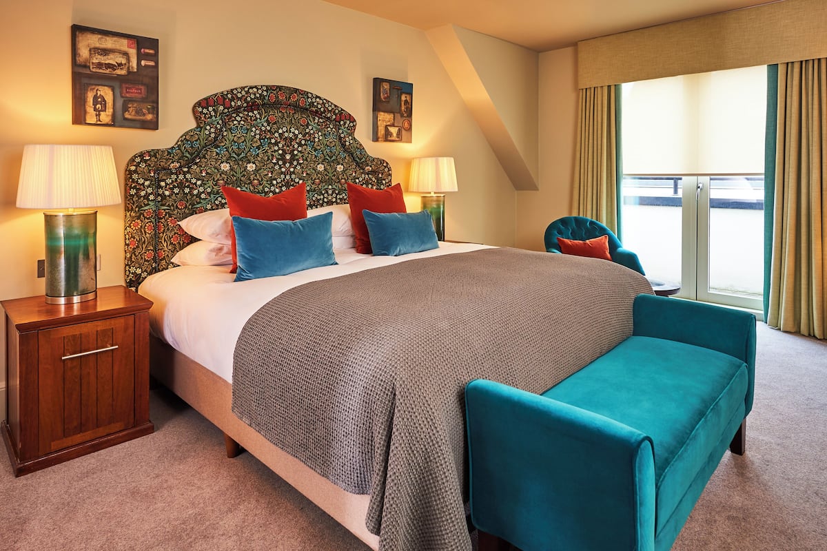 A hotel room with a large bed, two bedside tables, a teal armchair, and a view of the outside through a large window.
