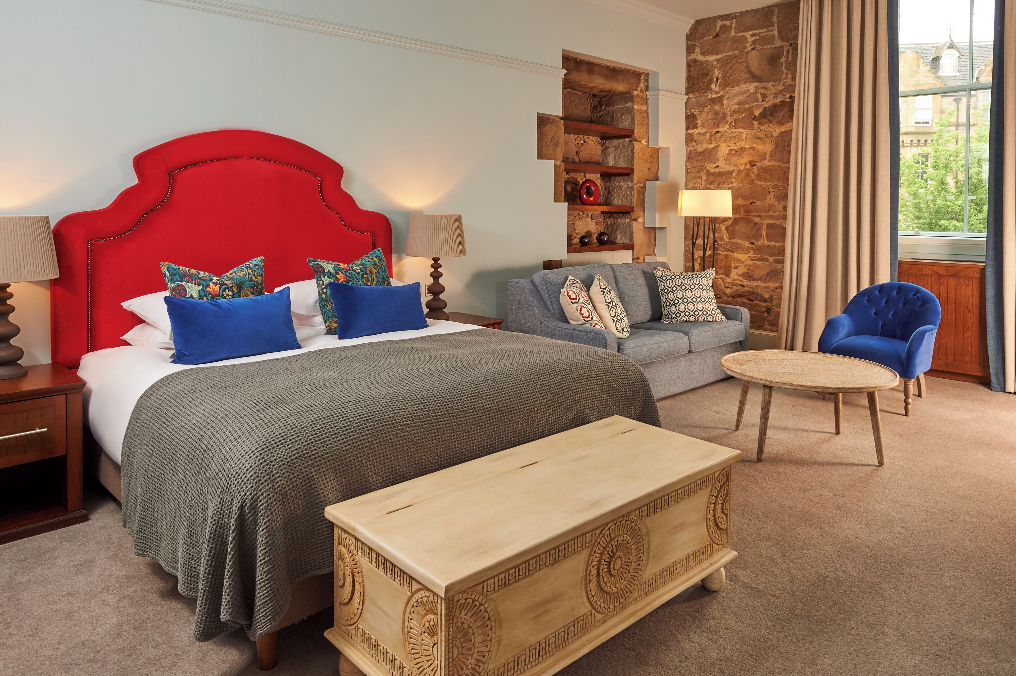 A bedroom with a red headboard, a grey sofa, a round wooden table, and a stone accent wall.