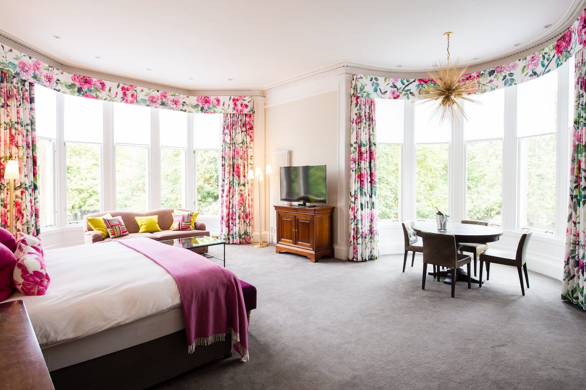 A bedroom with a large bed, a TV, a dining table with chairs, and floral curtains.