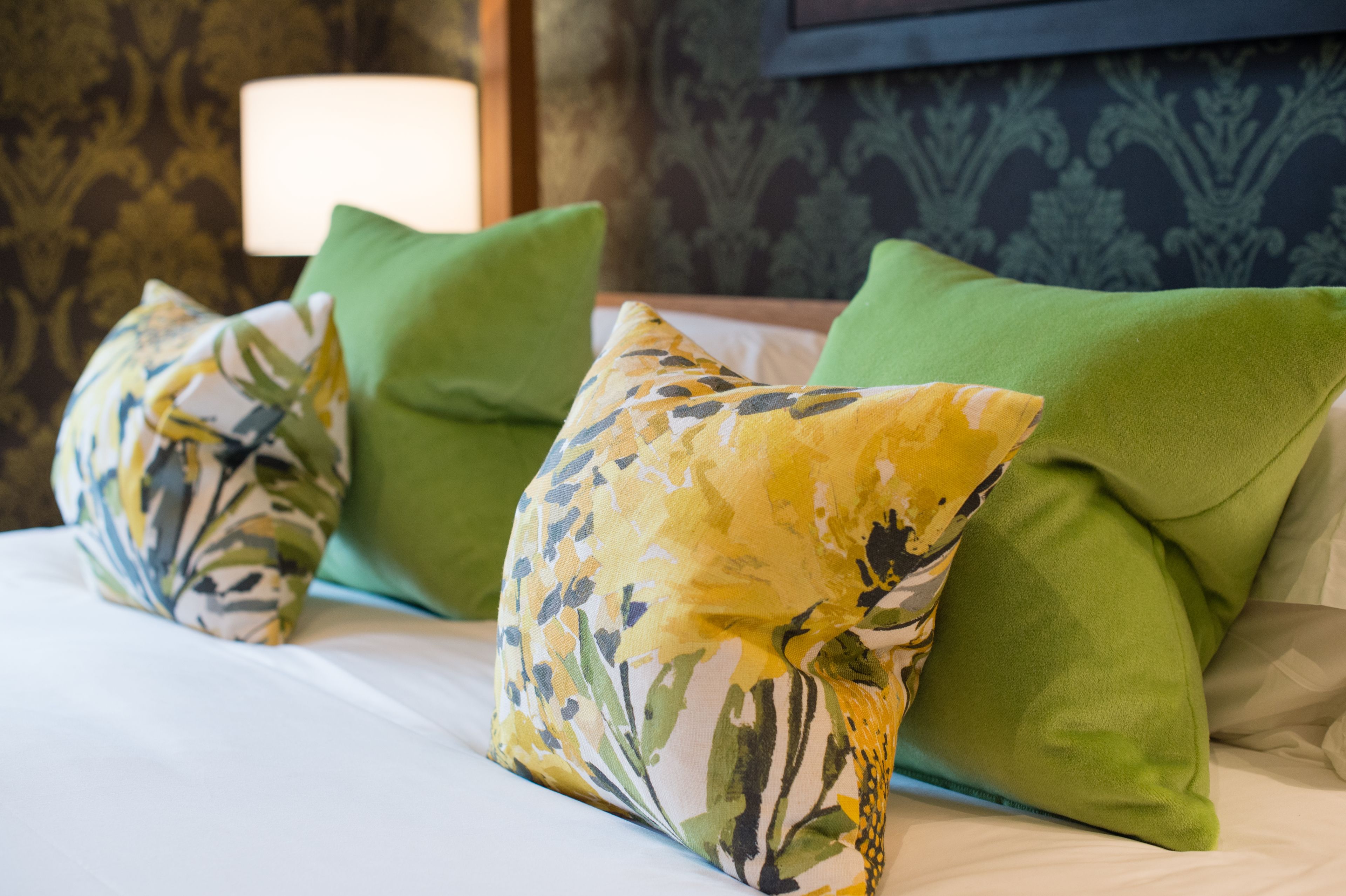 A bed with green and yellow patterned pillows, a white bedspread, and a wall with a patterned wallpaper and a framed picture.