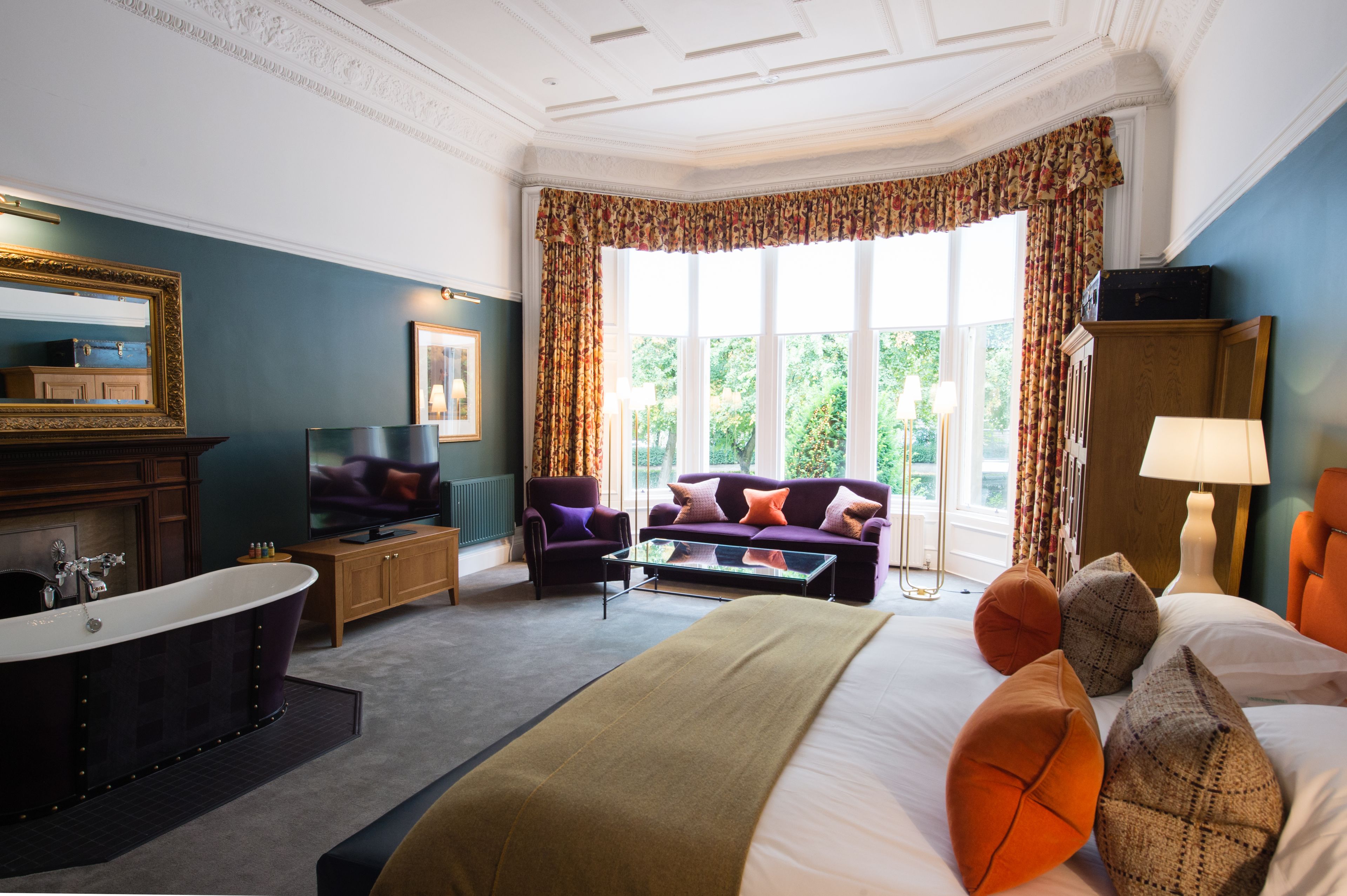 Quarto de hotel com uma cama grande, área de estar com sofá e poltronas, televisão, lareira e vista para o verde pelas janelas.