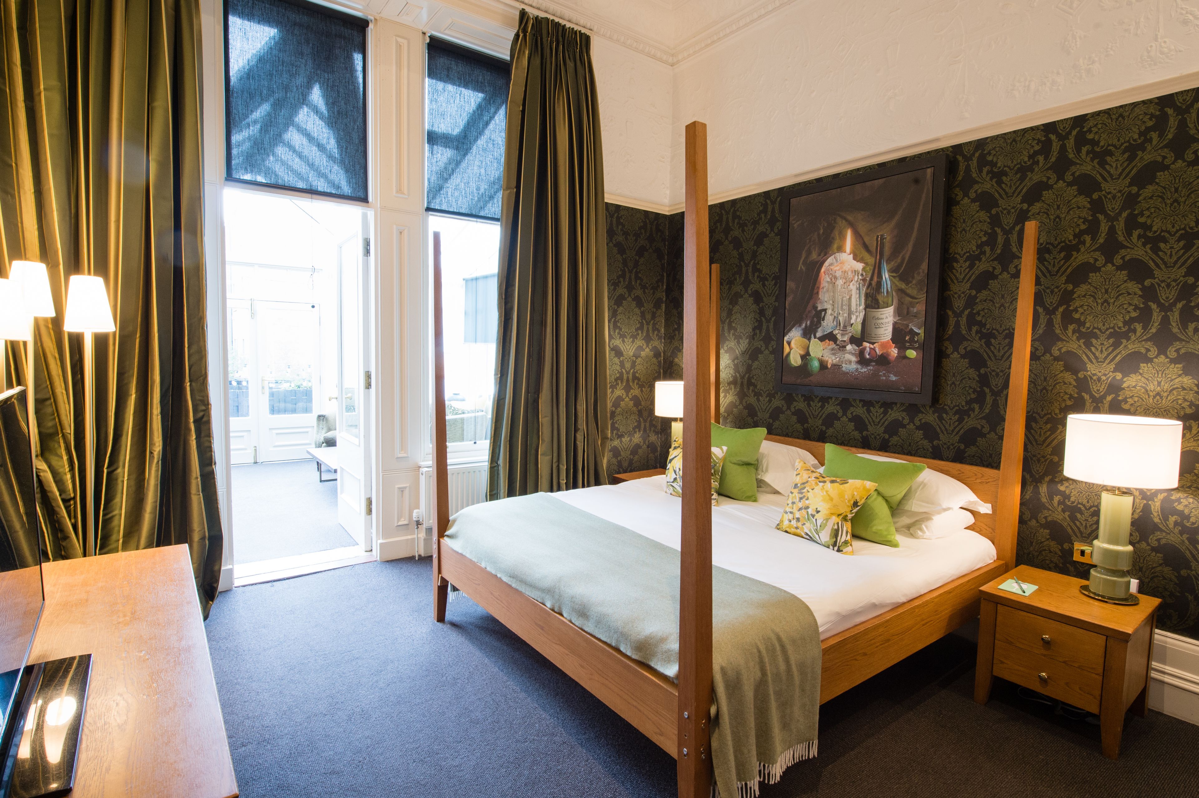 Une chambre d’hôtel avec un lit à baldaquin, une table de chevet avec une lampe, une vue sur l’extérieur à travers une porte vitrée et un tableau au mur.
