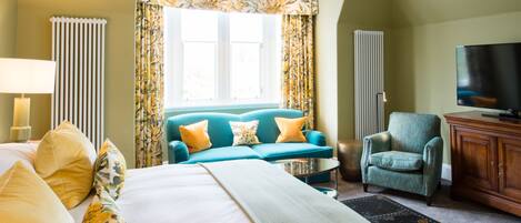 A hotel room with a bed, a blue sofa, a teal armchair, a television, and a window with curtains.