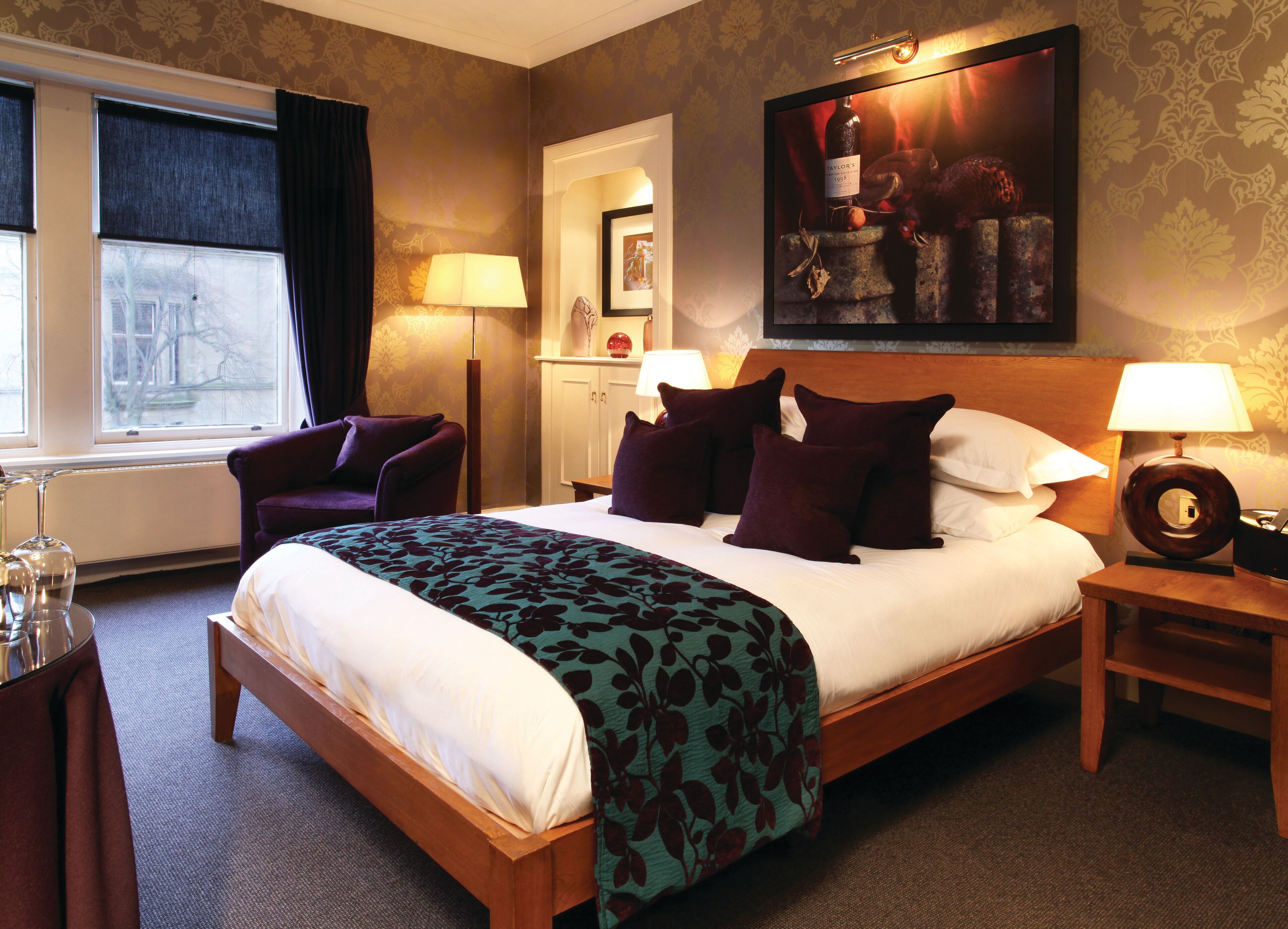 A hotel room with a large bed, a chair, a desk with a lamp, and a painting on the wall.