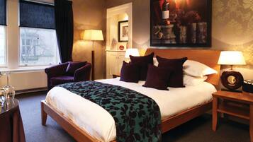 A hotel room with a large bed, a chair, a desk with a lamp, and a painting on the wall.
