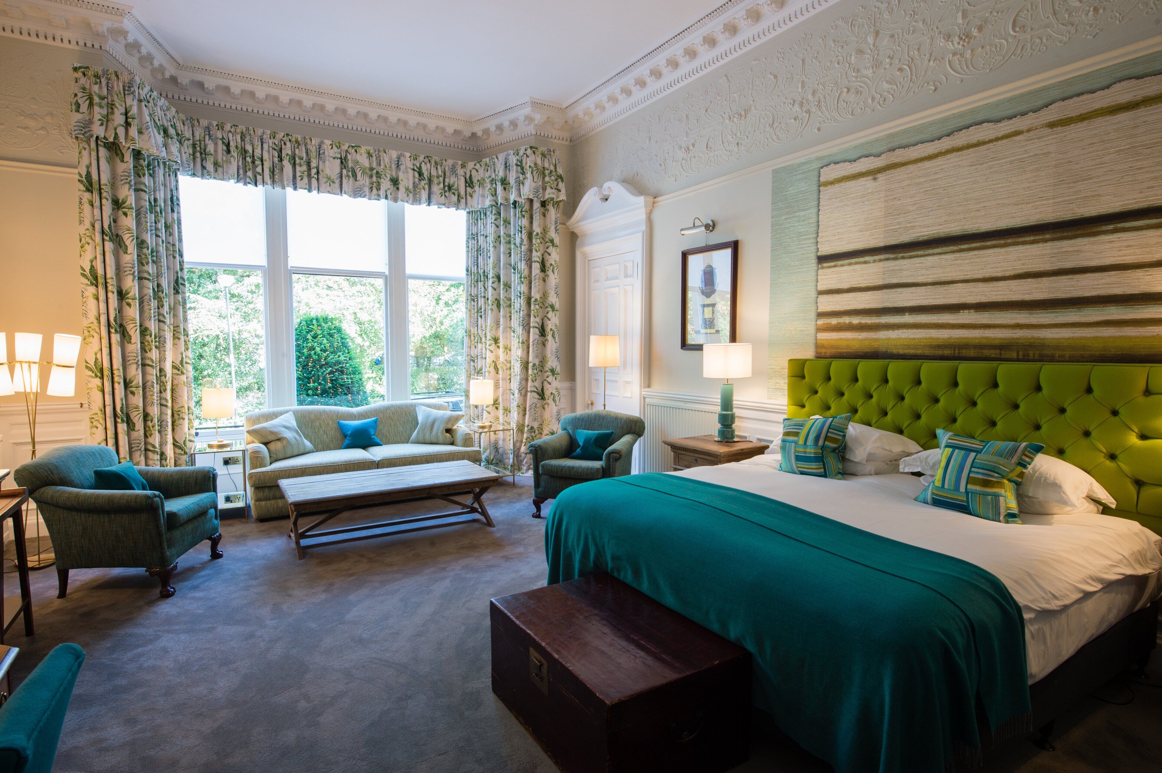 A bedroom with a green upholstered headboard, a large bed with multiple pillows, a green sofa, a small table, and a chair.