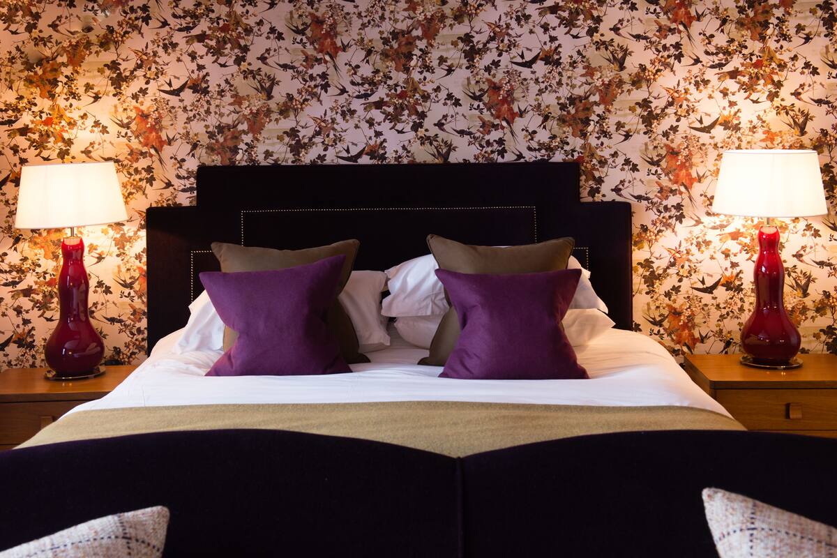 A neatly made bed with purple pillows, two red lamps on either side, and a floral wallpaper background.