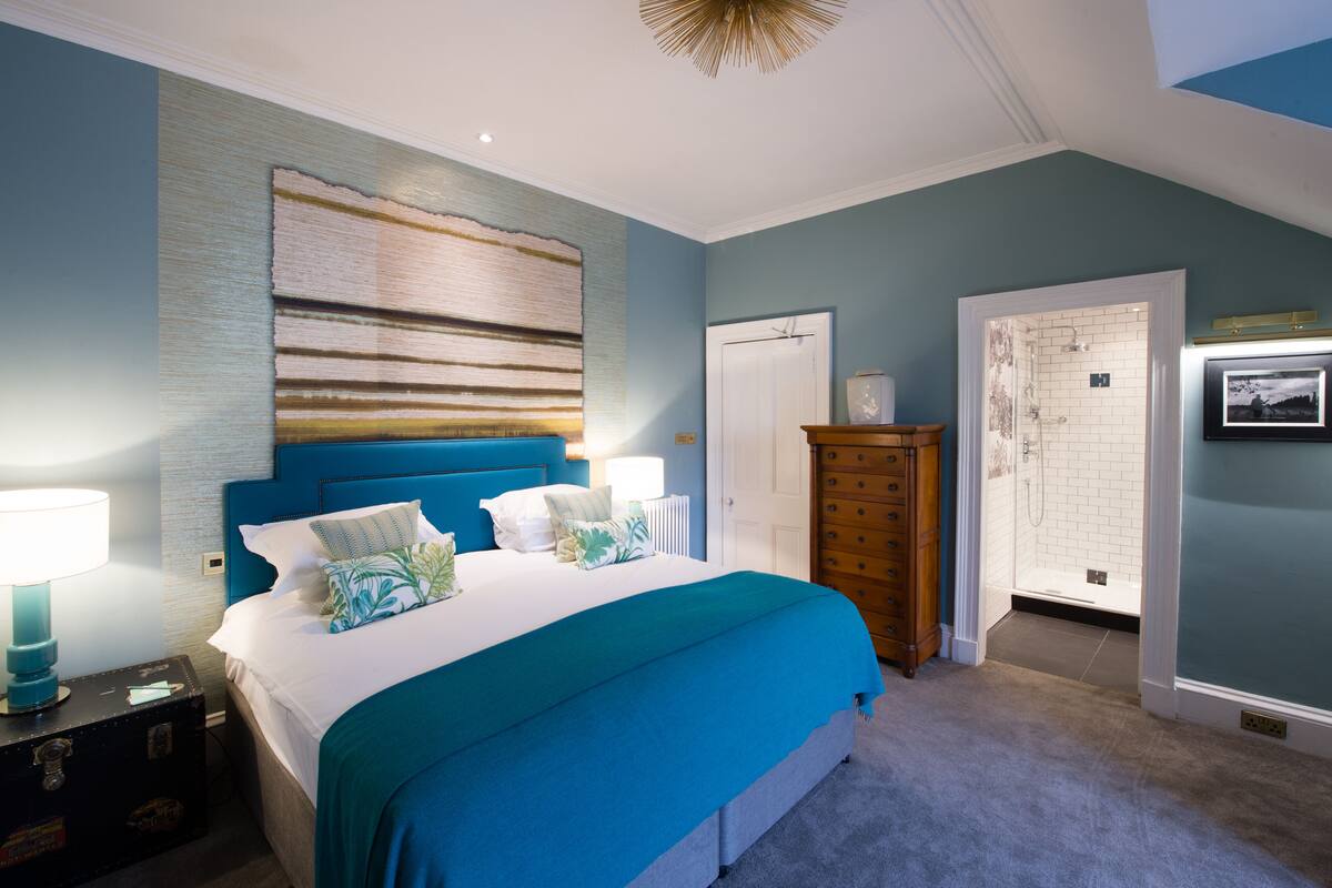 A bedroom with a large bed, a dresser, a nightstand, and a bathroom with a shower and toilet visible.