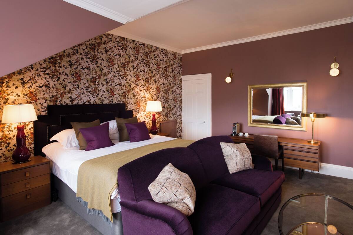 A bedroom with a large bed, two bedside tables with lamps, a dresser, a mirror, and a purple sofa.