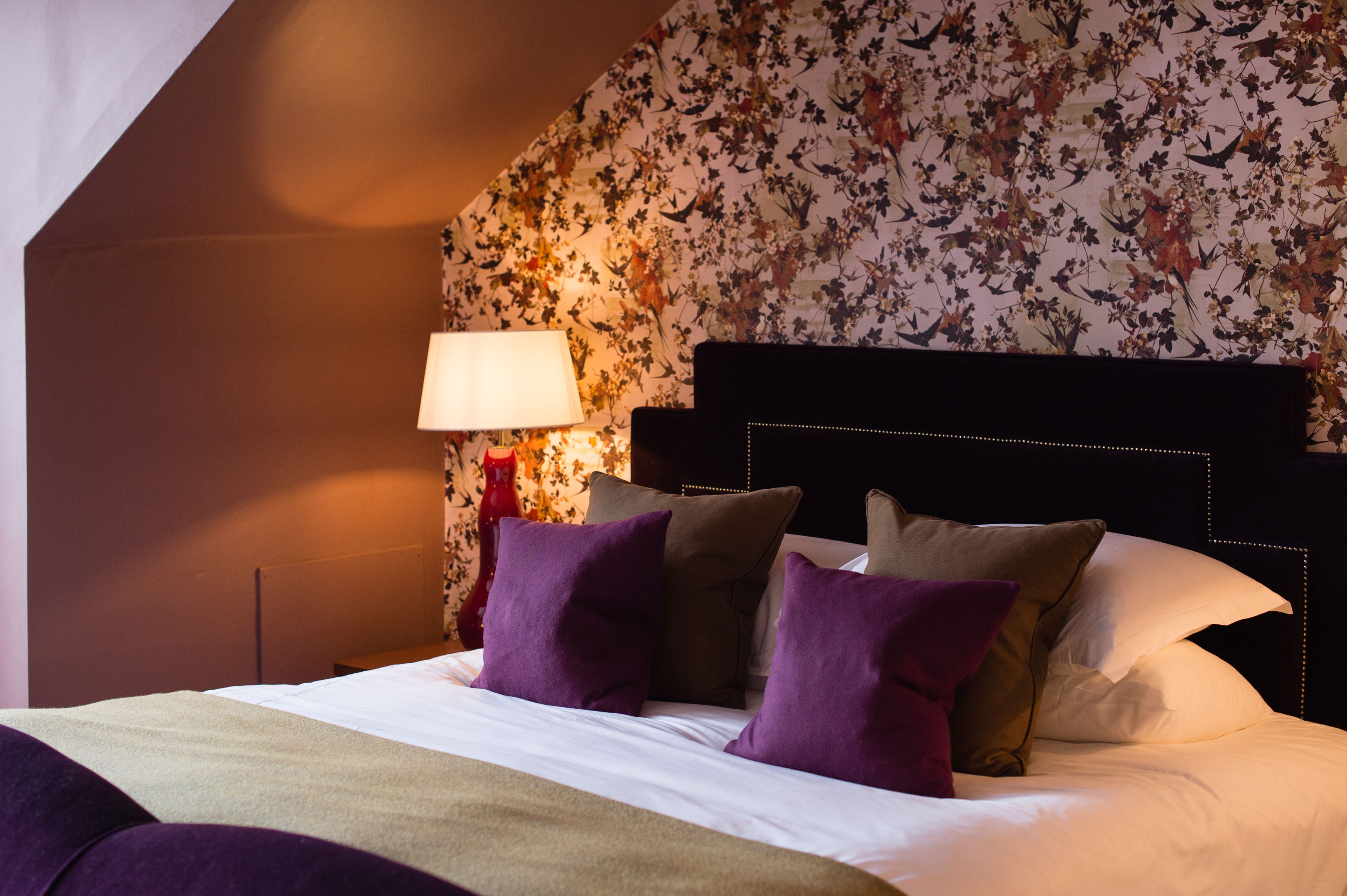 Een hotelkamer met bloemenbehang, een zwart hoofdeinde en een bed met meerdere kussens.