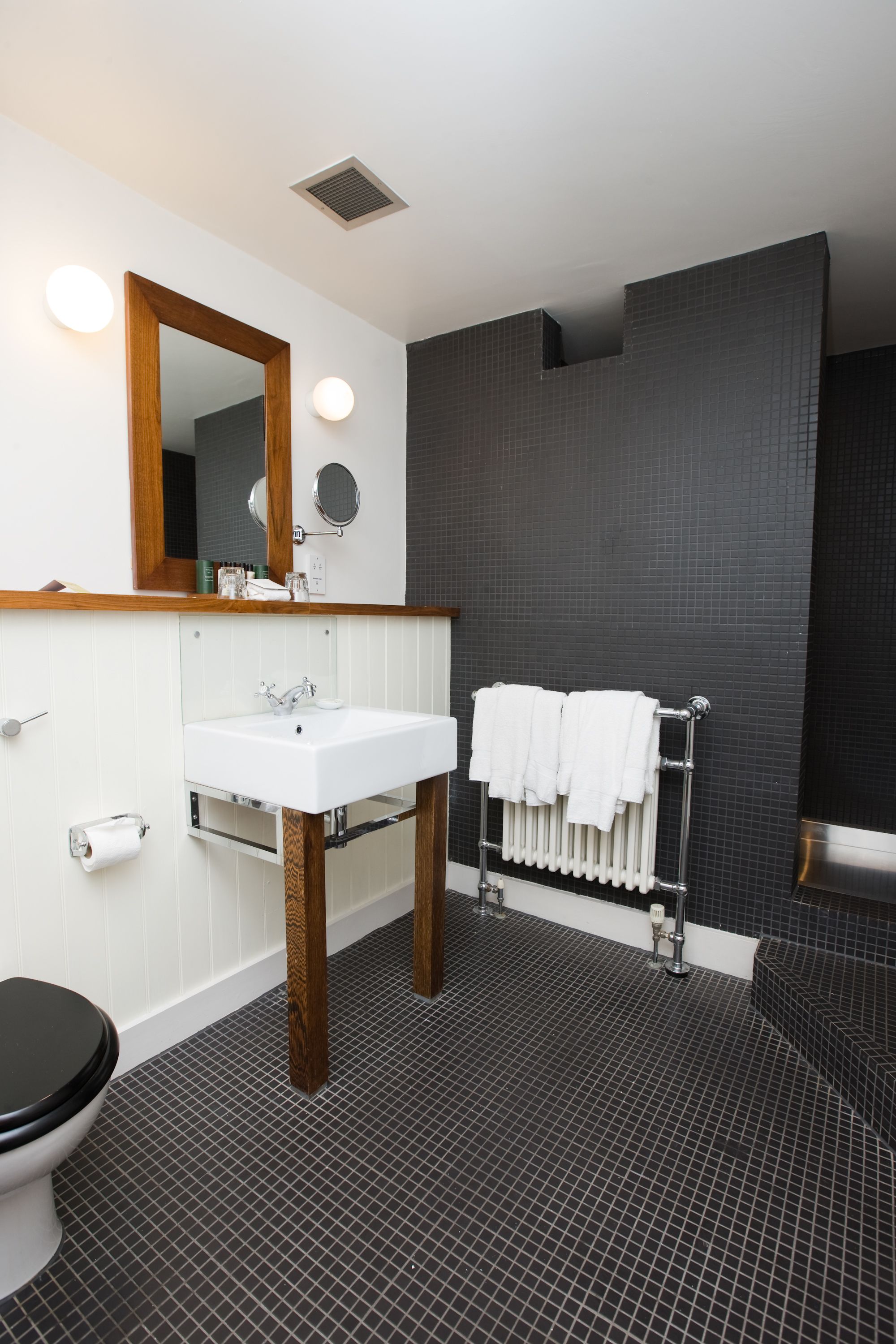 A modern bathroom with a white sink, wooden vanity, a large mirror, a towel rack, and a black tiled floor.
