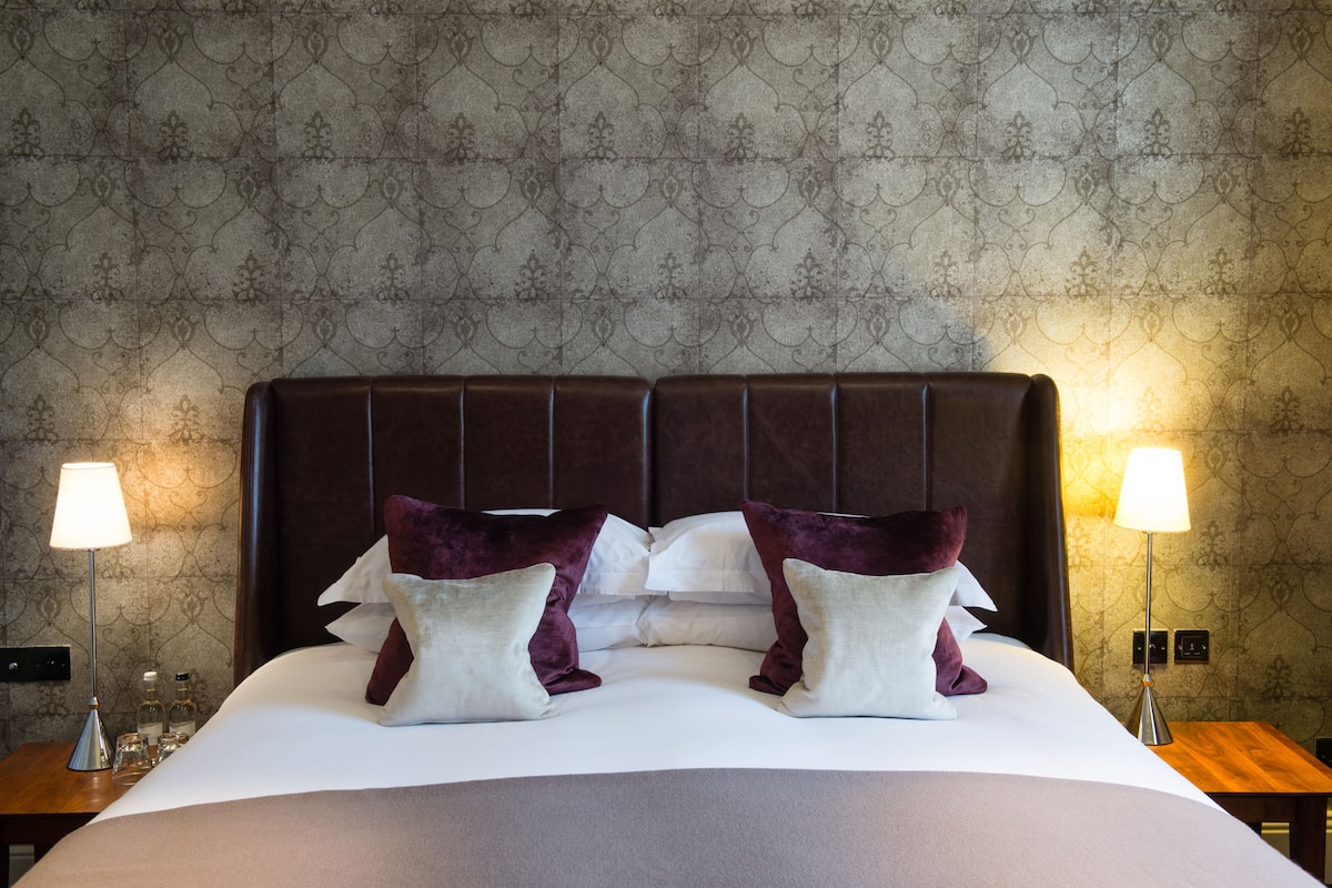 A neatly made bed with a dark headboard, white linens, and two decorative pillows. There are two bedside lamps on wooden nightstands.