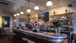 Zona de bar con una barra larga, varios taburetes, luces colgantes y estantes llenos de botellas.