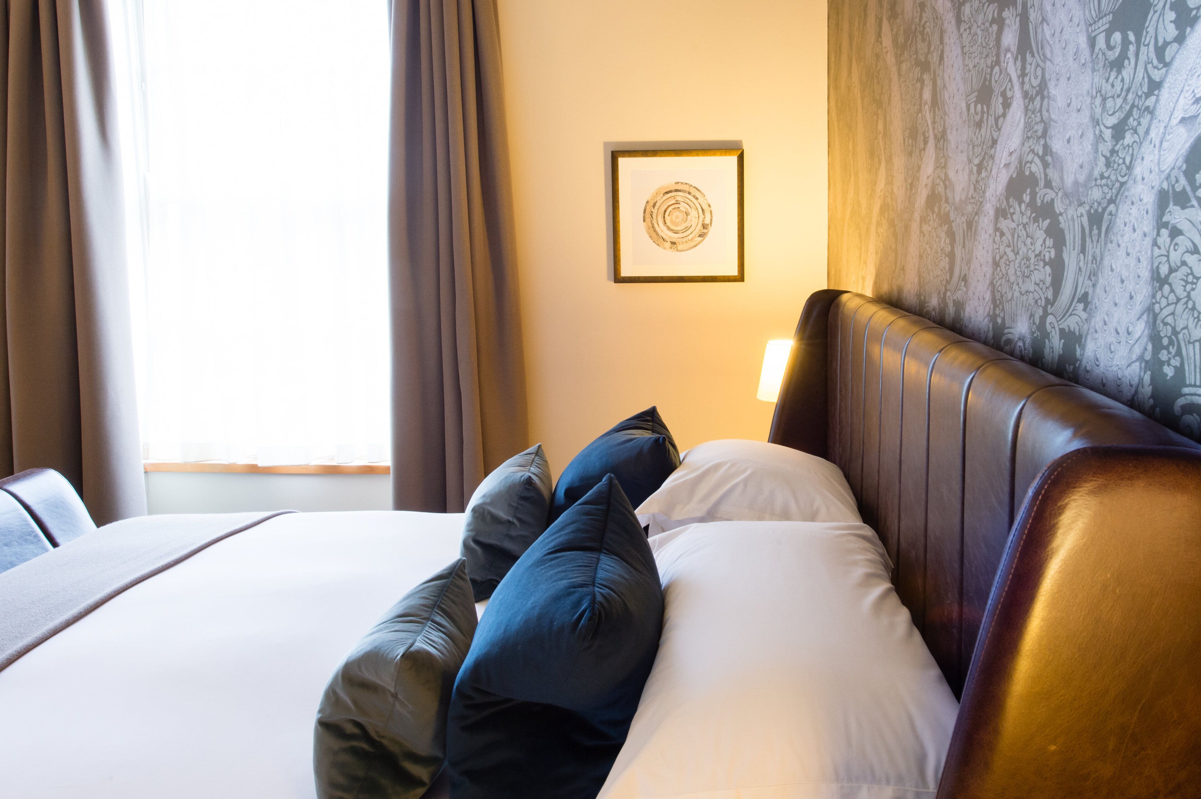 A hotel room with a bed, pillows, a headboard, a wall with a framed picture, and curtains.