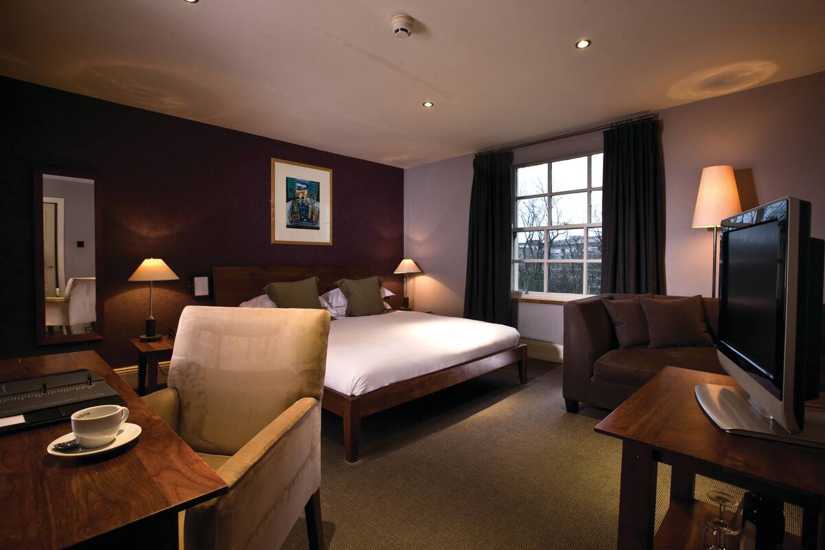 A hotel room with a bed, two chairs, a desk with a laptop, a television, and a window with curtains.