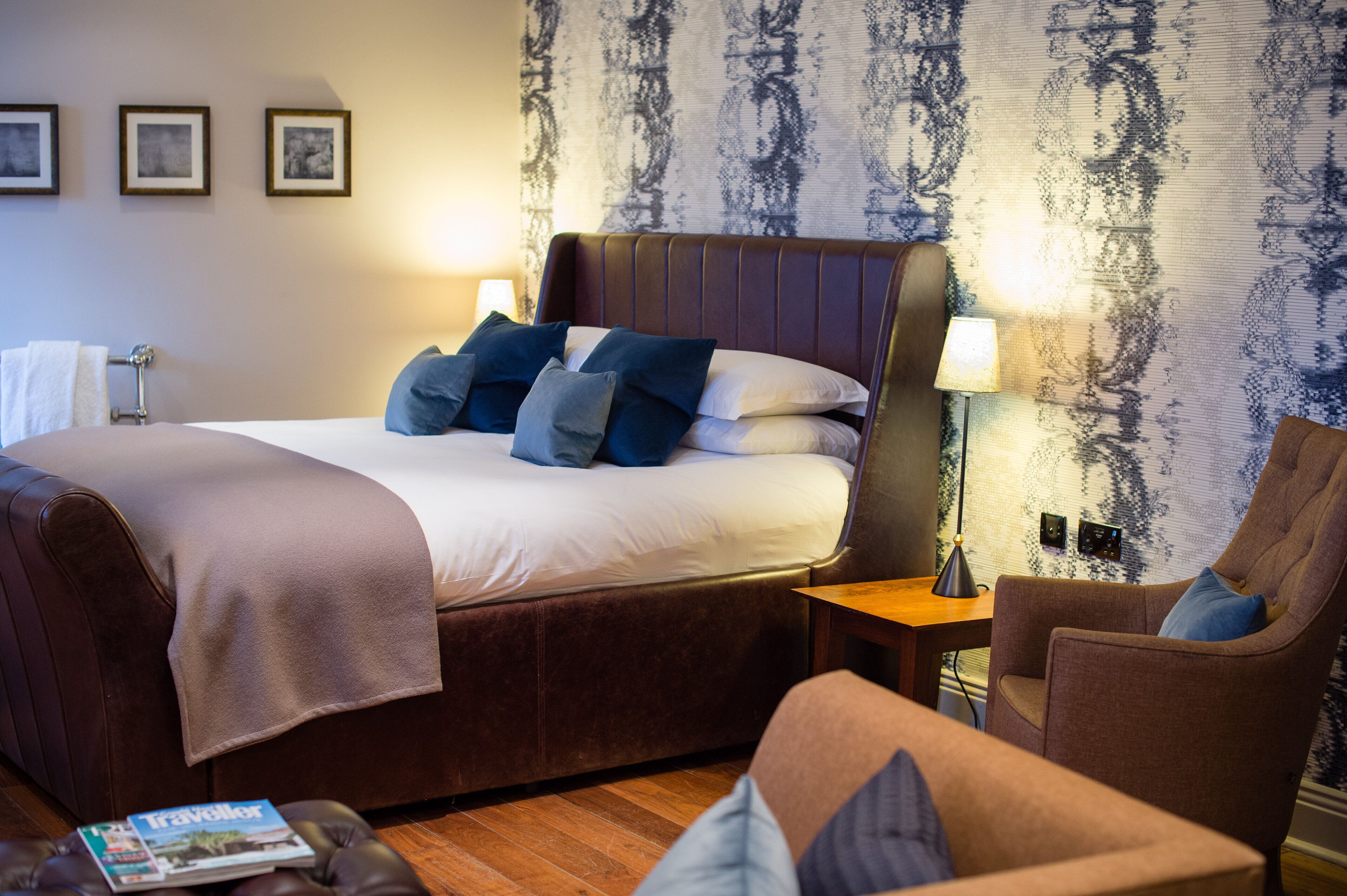 Habitación de hotel con una cama grande, una mesita de noche de madera, un sillón marrón y una revista en el suelo.