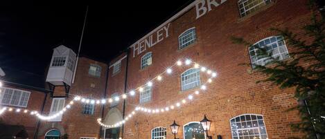 Un edificio de ladrillo con el nombre "HENLEY BREWER" en la fachada, mesas y sillas para asientos al aire libre y luces colgantes.