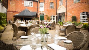 An outdoor dining area with wicker tables and chairs, a brick building in the background, and string lights.