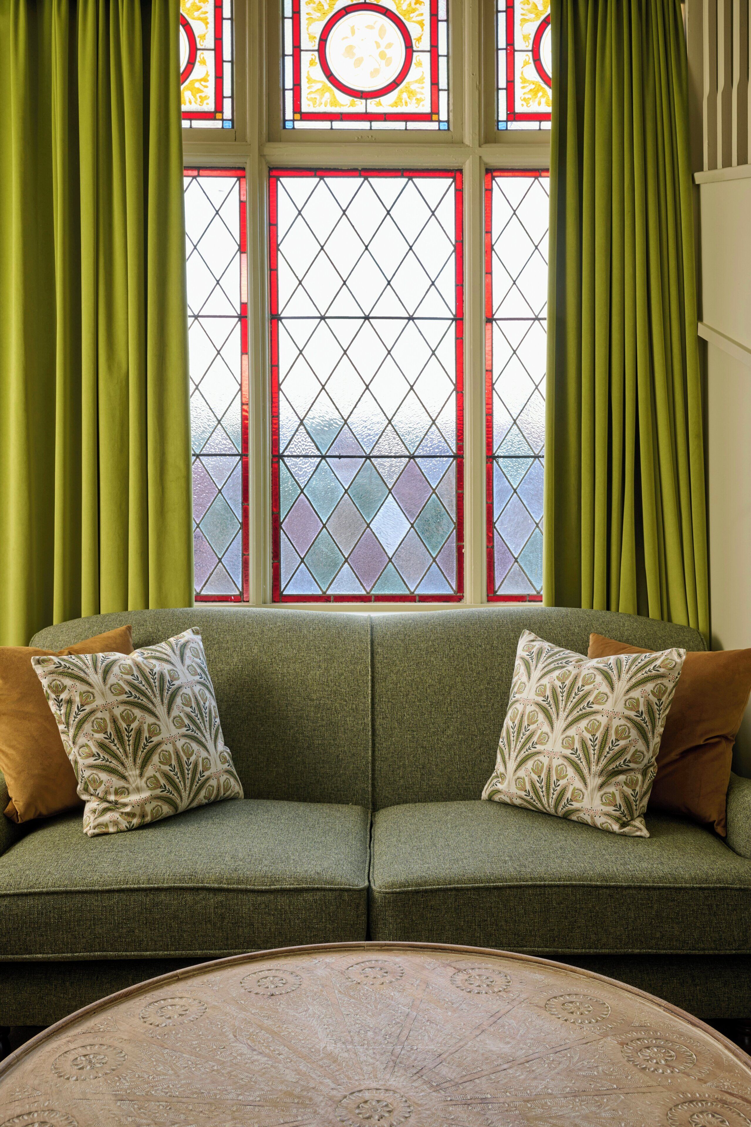 A green sofa with patterned cushions, a round table, and a window with green curtains and stained glass.