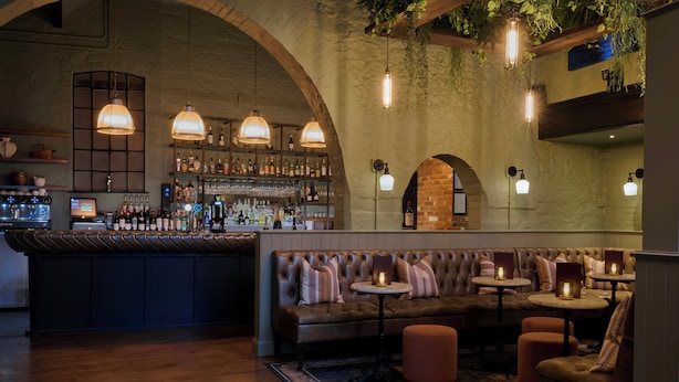 A bar area with a long counter, multiple seating options including a sofa and round tables, and hanging pendant lights.