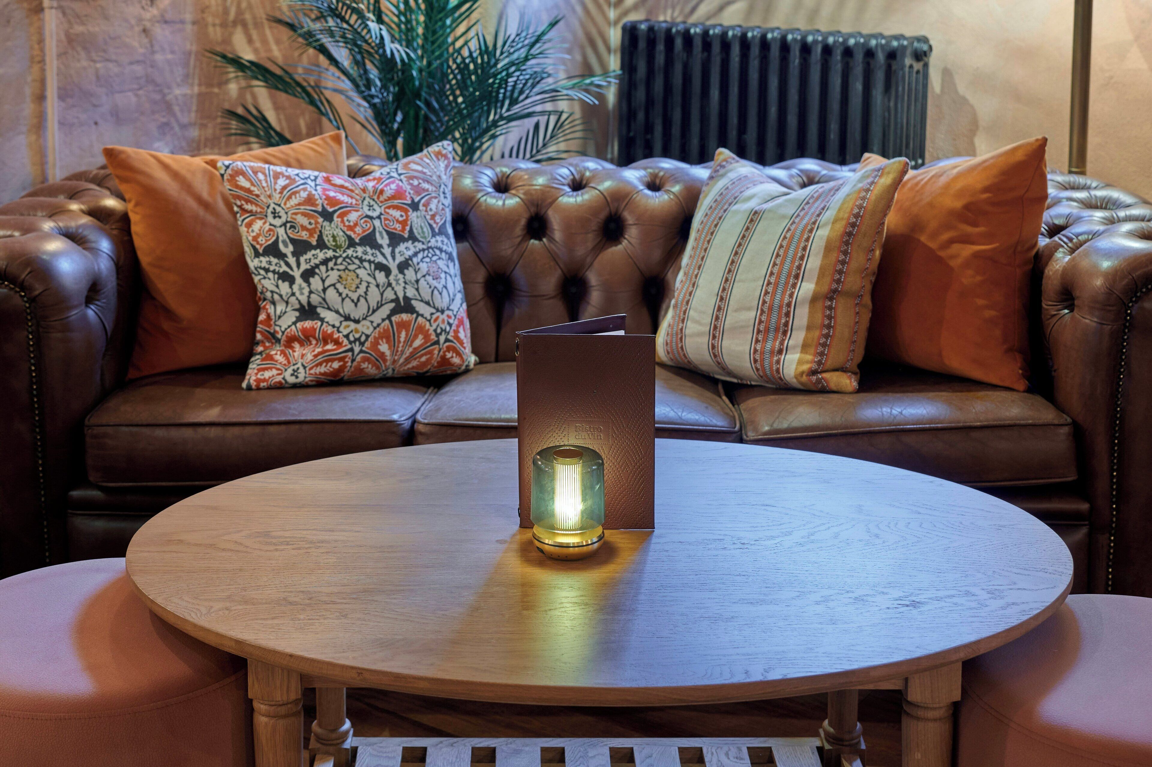 A leather sofa with floral cushions, a round wooden table with a candle, and a radiator in the background.