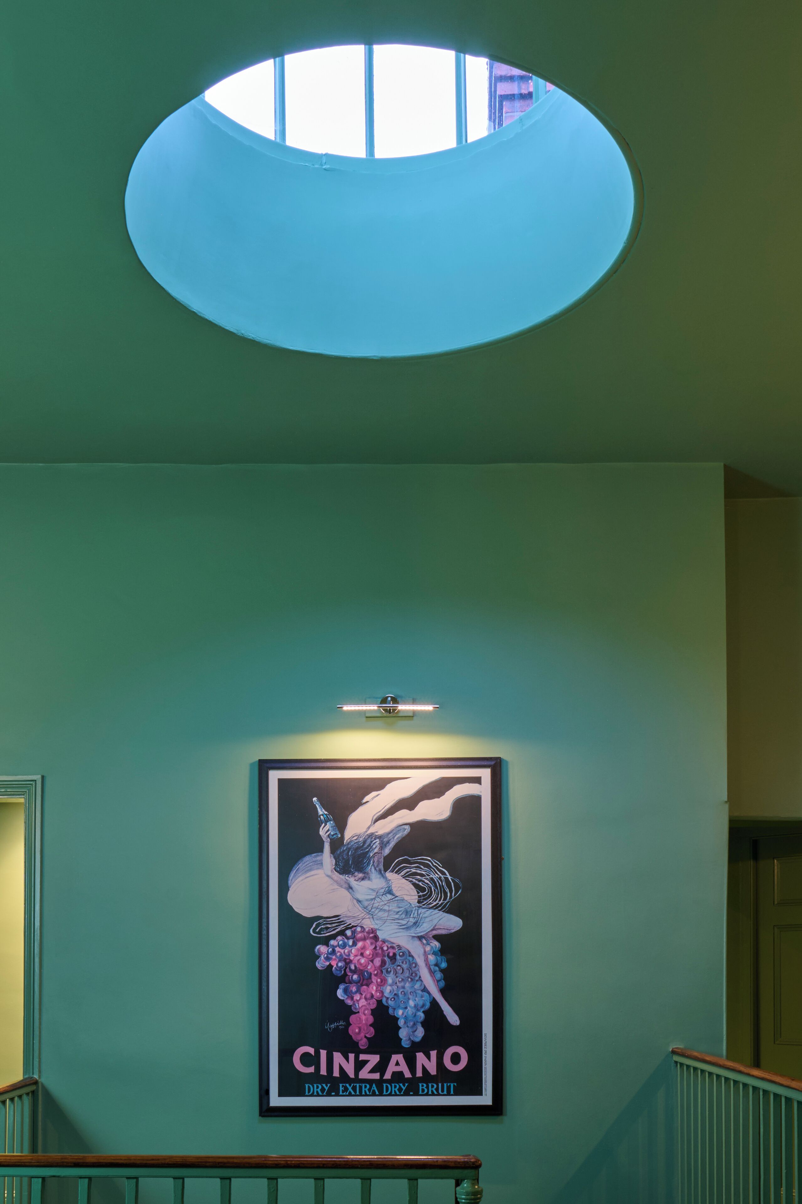 A framed poster of a woman with grapes on a teal wall, a skylight, and a staircase with wooden railings.