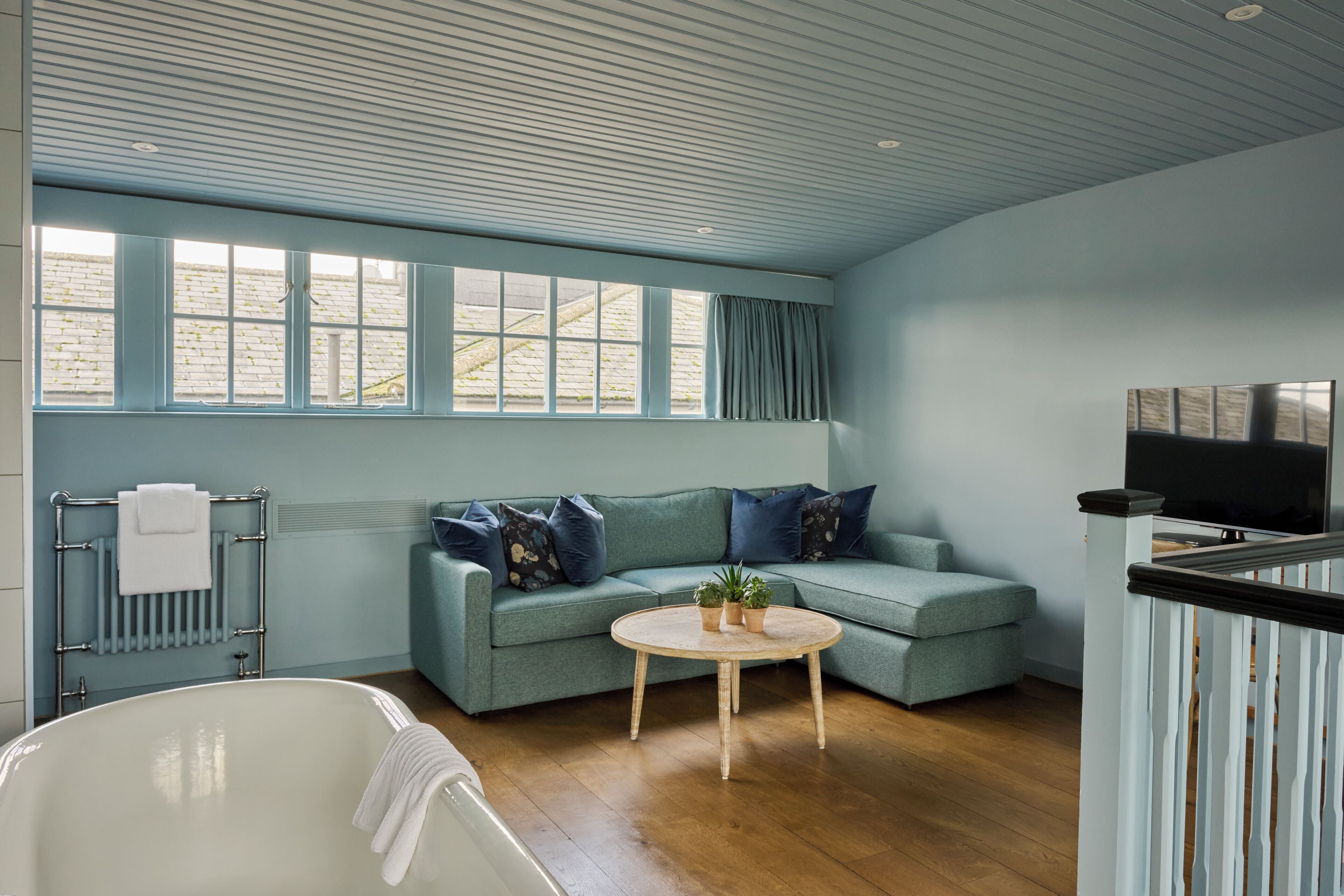 Una habitación con suelo de madera, un sofá azul claro con cojines oscuros, una mesa de centro redonda de madera con dos plantas en macetas, una bañera blanca y un amplio ventanal con cortinas.