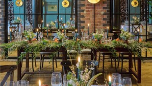 A restaurant with a long table adorned with greenery and candles, set against a brick wall with large windows.