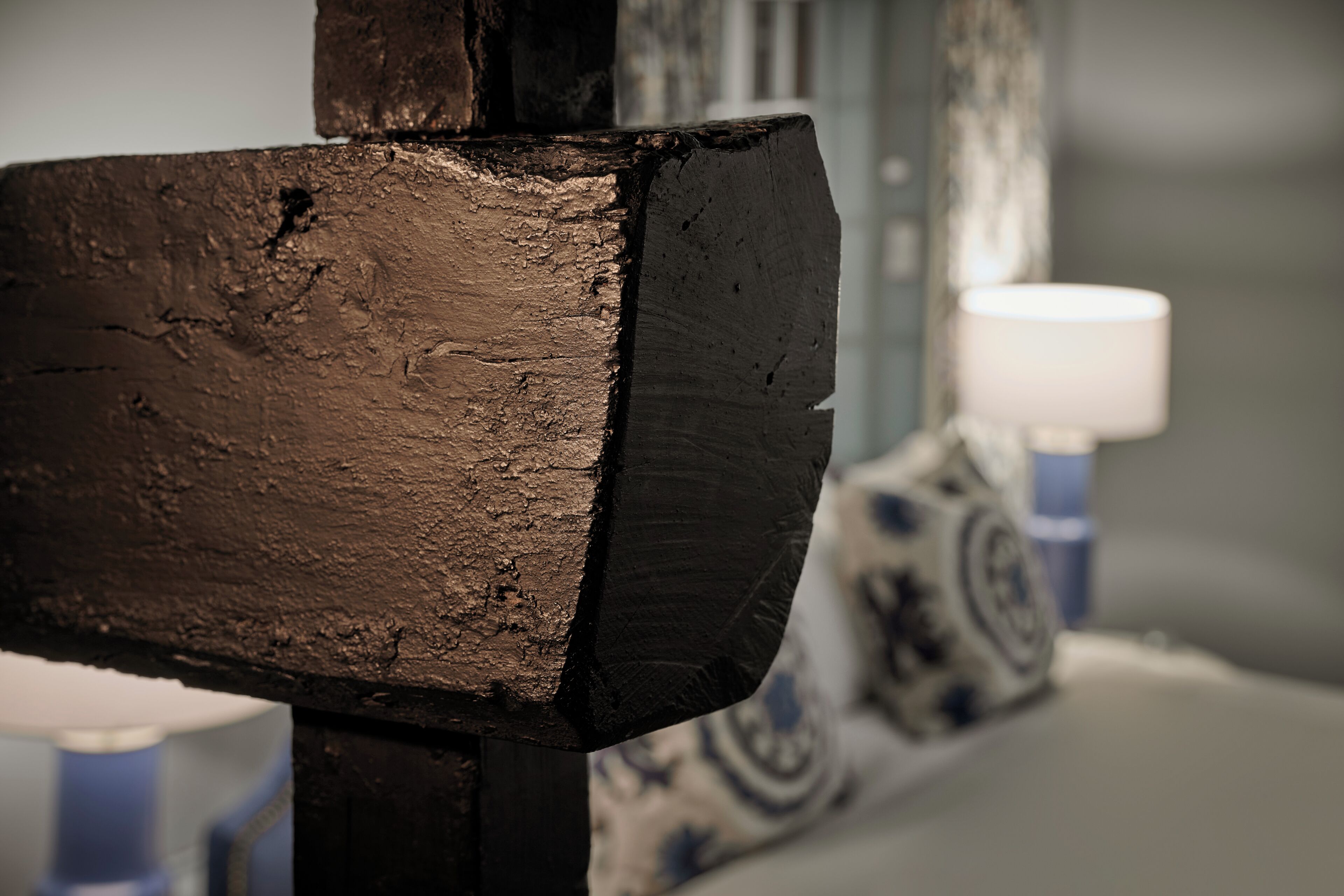 A close-up of a wooden chair with a textured surface, a lamp in the background, and a patterned wall.