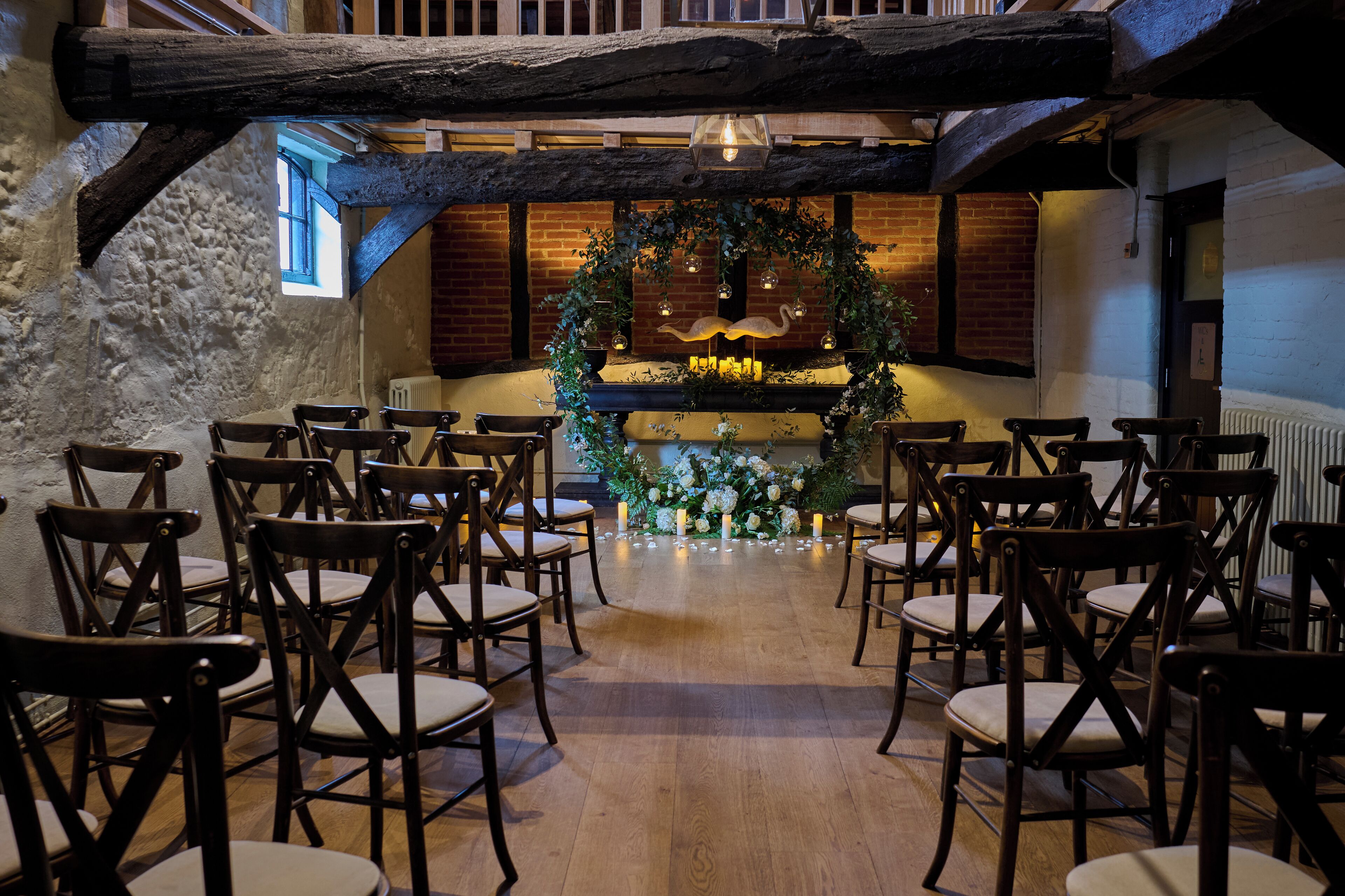 A rustic interior with wooden chairs arranged in rows, a floral arch, and a brick wall with a window.