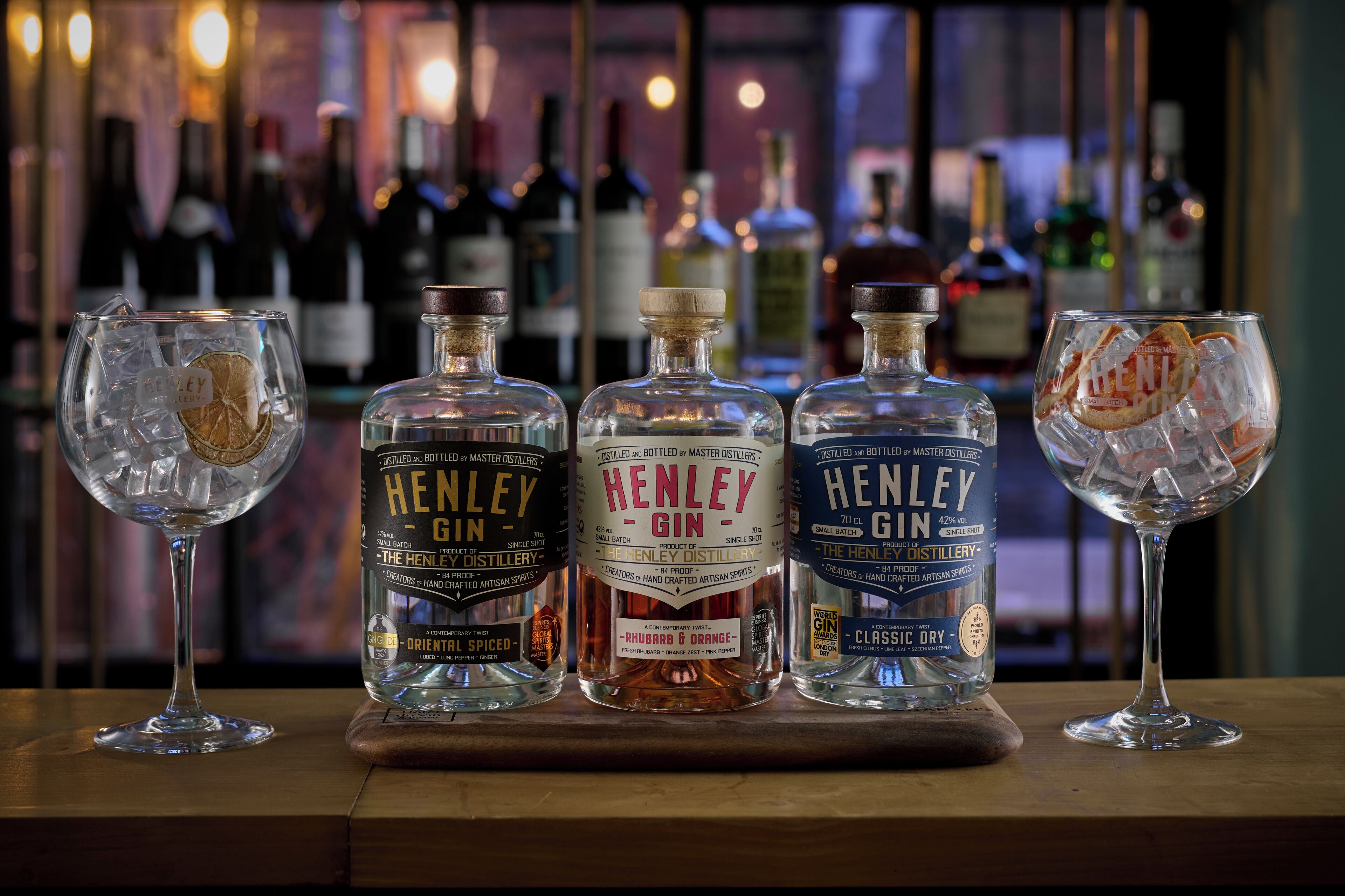 Three bottles of Henley gin and two glasses on a wooden surface with a blurred background of bottles.