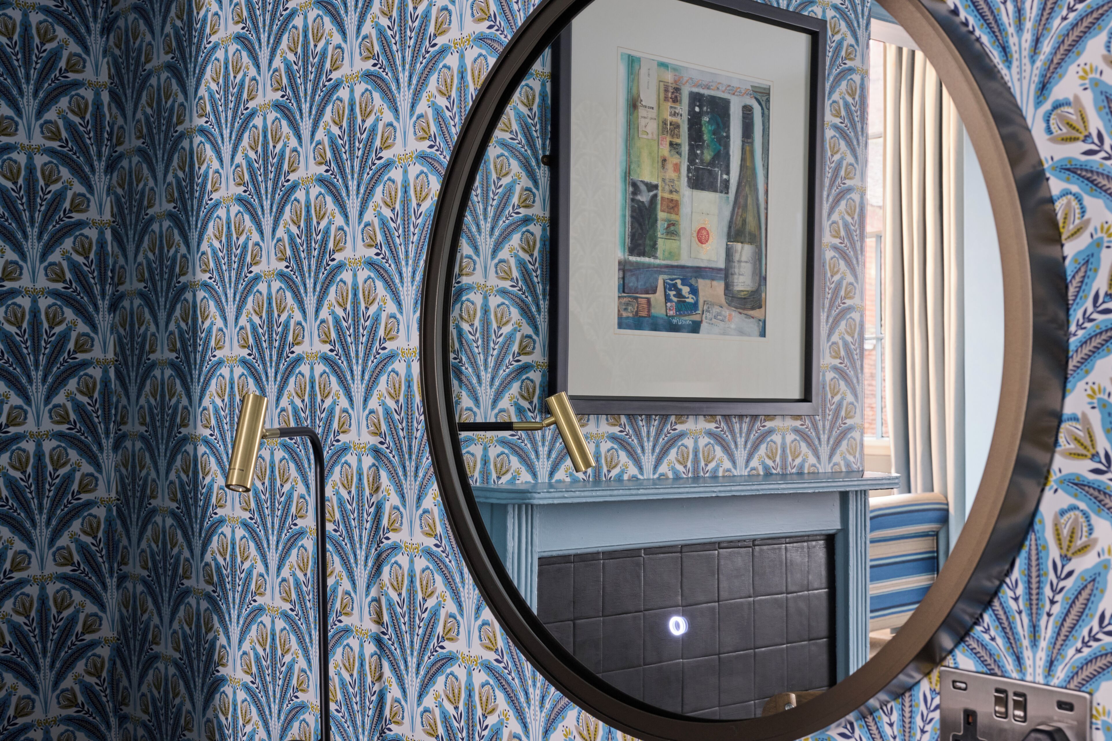 A room with a patterned wallpaper, a framed artwork on the wall, and a round mirror reflecting part of the room.