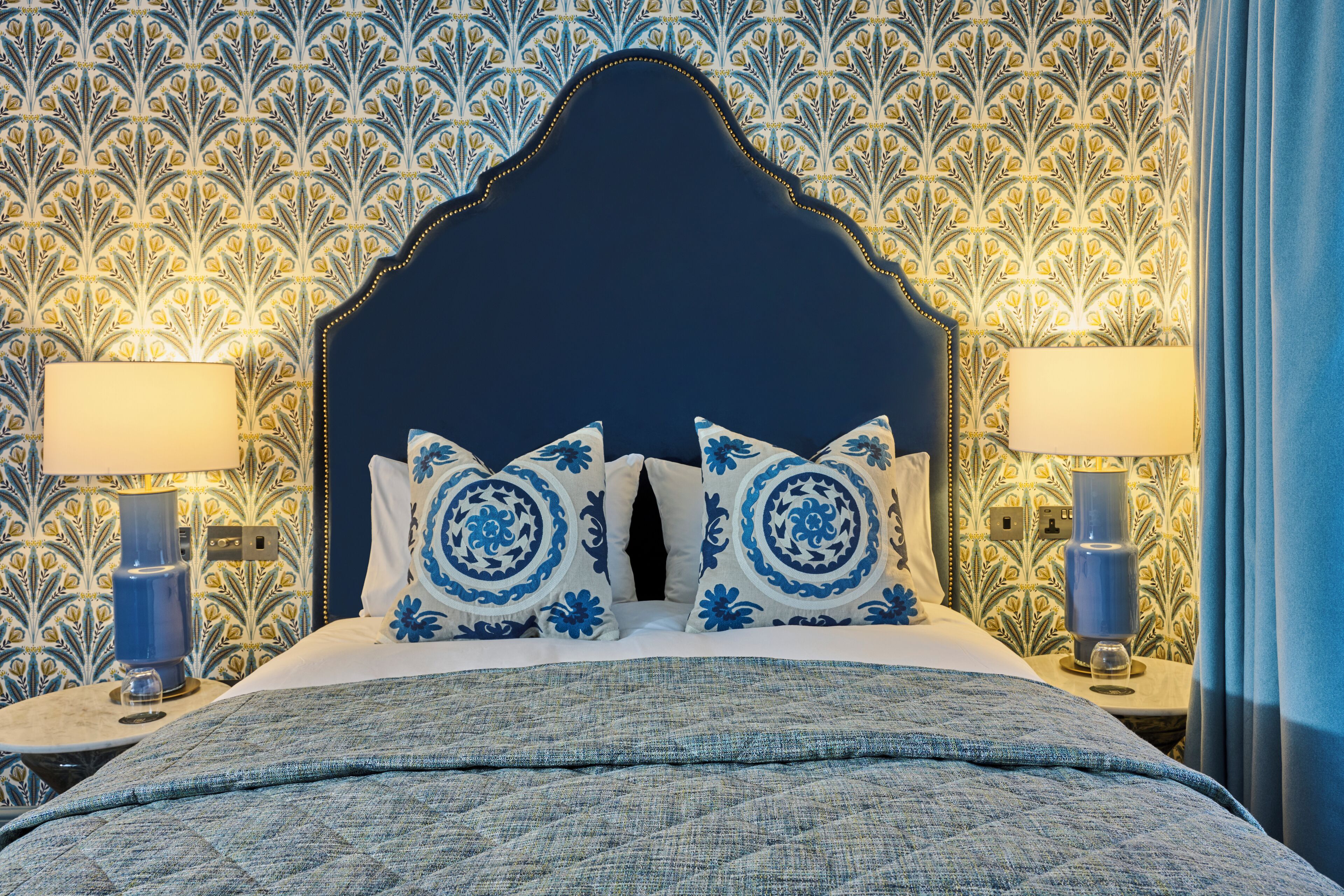 Una cama bien hecha con cabecera azul, dos lámparas a juego y papel tapiz estampado.
