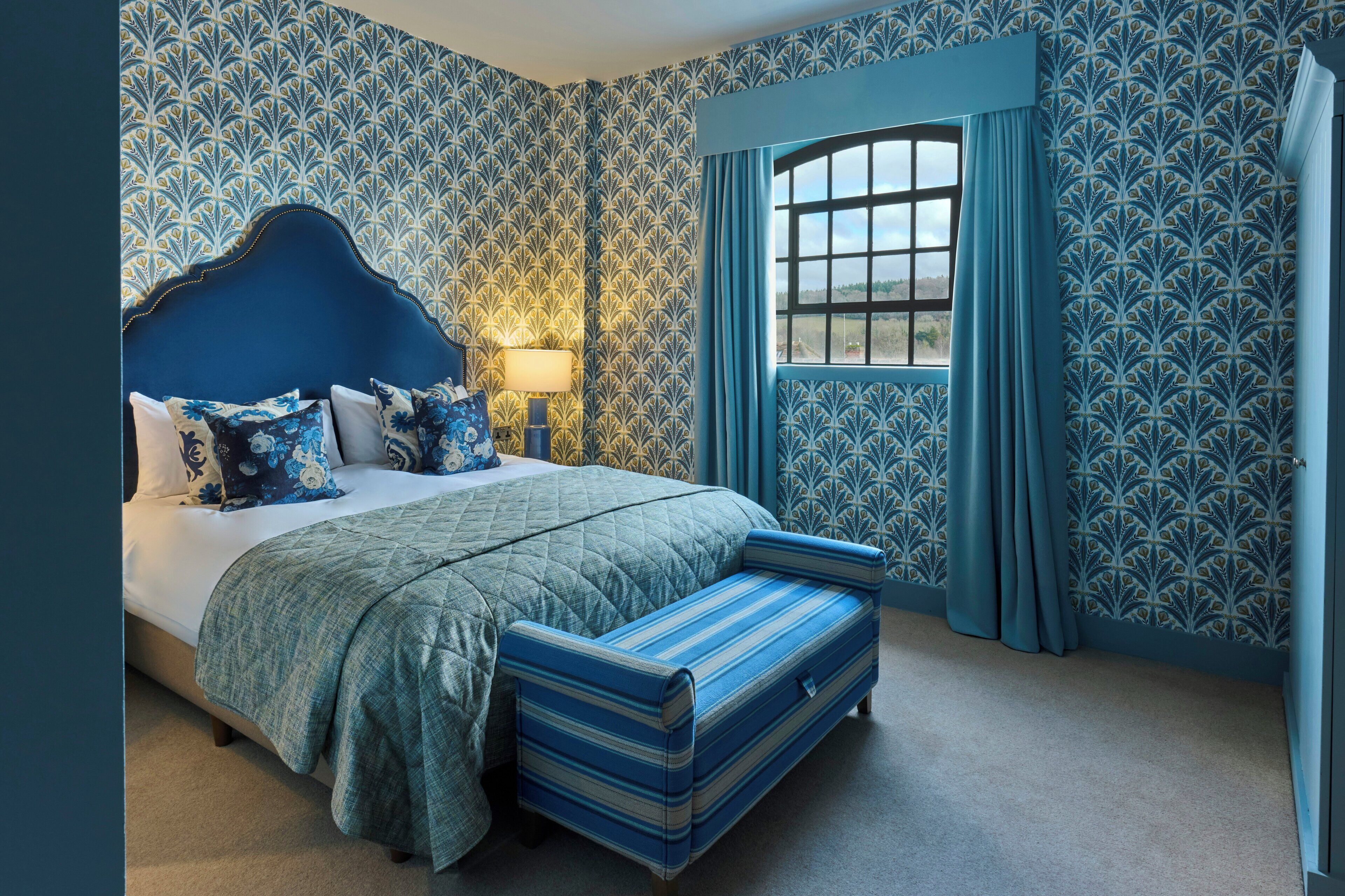 A bedroom with a large bed, a nightstand, a window with curtains, and patterned wallpaper.